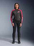 Frau in Alpinestars Stella T-Dyno WR Jacke, Schwarz mit leuchtend roten und fuchsiafarbenen Akzenten, Textiljacke im Vintage-Stil mit pinkfarbenem Logo und Ärmelstreifen, kombiniert mit schwarzer Hose und Schnürstiefeln, urbane Motorradausrüstung