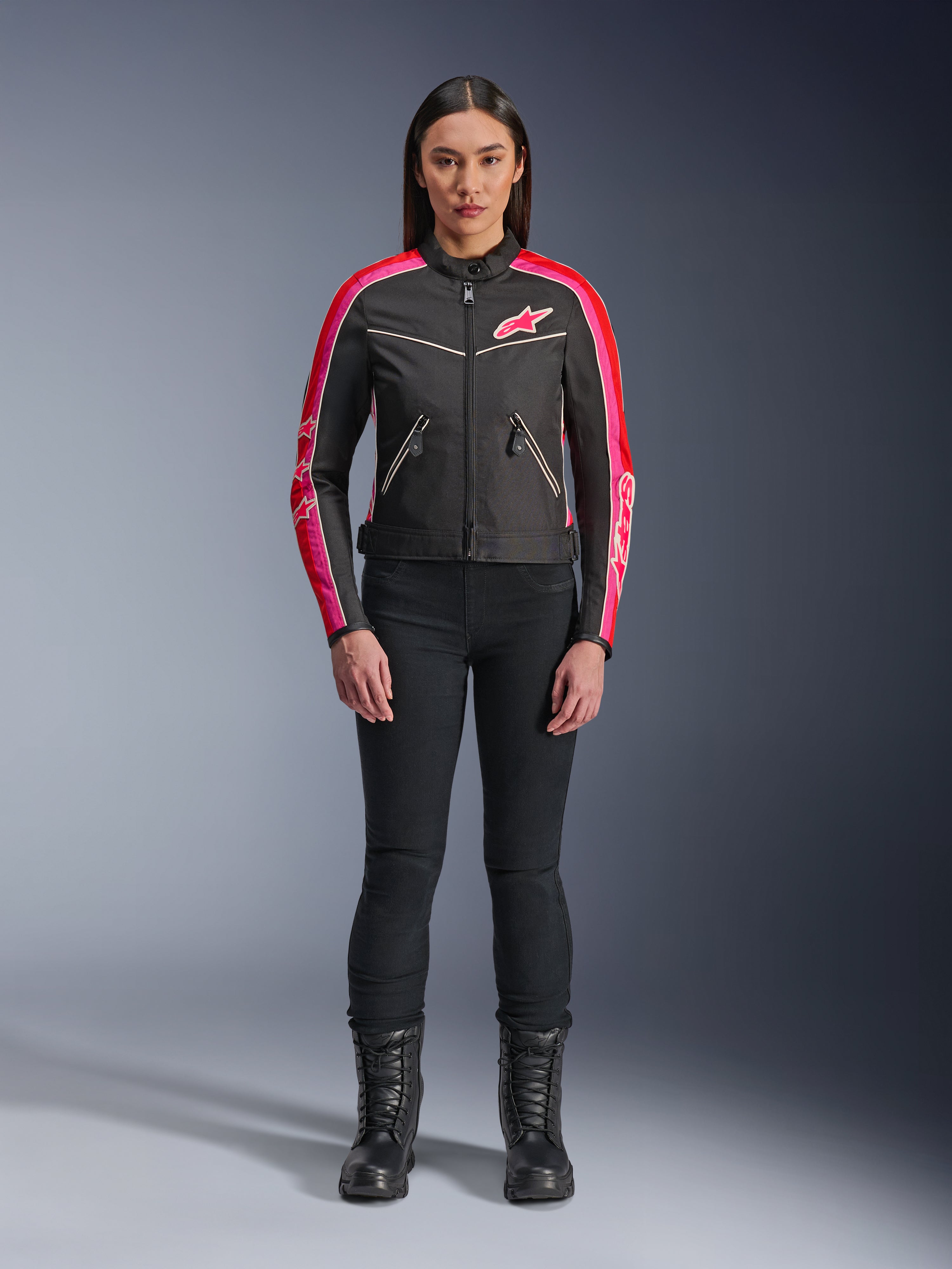 Frau in Alpinestars Stella T-Dyno WR Jacke, Schwarz mit leuchtend roten und fuchsiafarbenen Akzenten, Textiljacke im Vintage-Stil mit pinkfarbenem Logo und Ärmelstreifen, kombiniert mit schwarzer Hose und Schnürstiefeln, urbane Motorradausrüstung