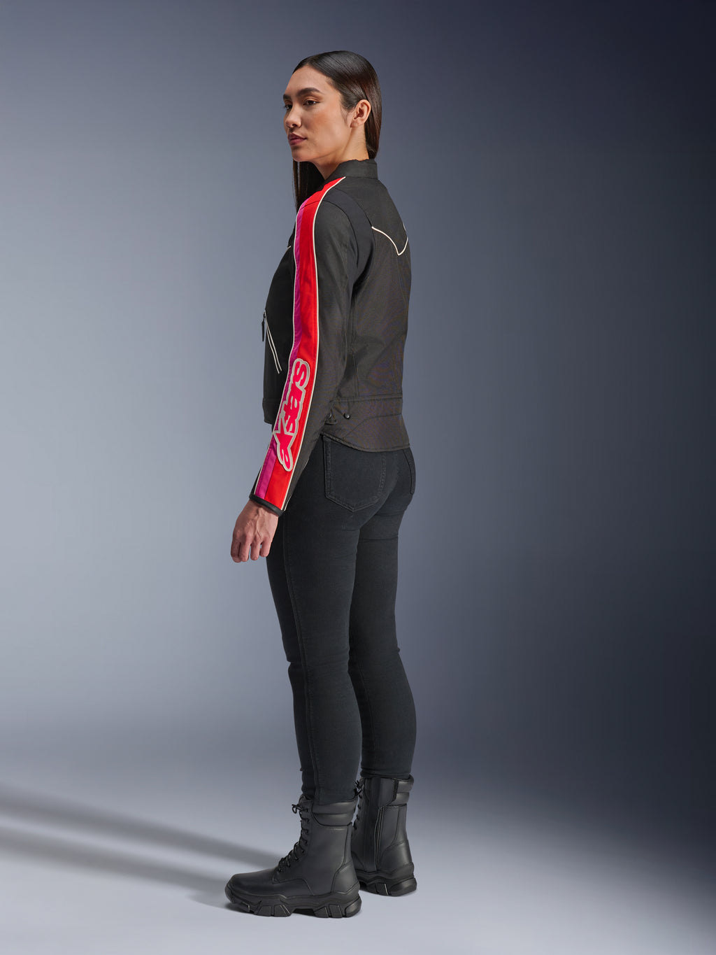 Weibliches Model trägt die Alpinestars Stella T-Dyno WR Jacke in Schwarz Bright Red Fuchsia, mit markanten roten und pinken Streifen und Alpinestars-Branding auf dem Ärmel, kombiniert mit schwarzer Motorradhose und schwarzen Motorradstiefeln in einer Studioumgebung.