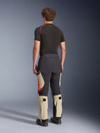 Alpinestars Halo Pro Drystar® XF Laminierte Hose, Adventure-Motorradhose, Mastic Black und Tan mit orangefarbenen Akzenten, Rückansicht zeigt die 3-lagige laminierte Schalenkonstruktion, kombiniert mit einem schwarzen Base-Layer und Adventure-Stiefeln.