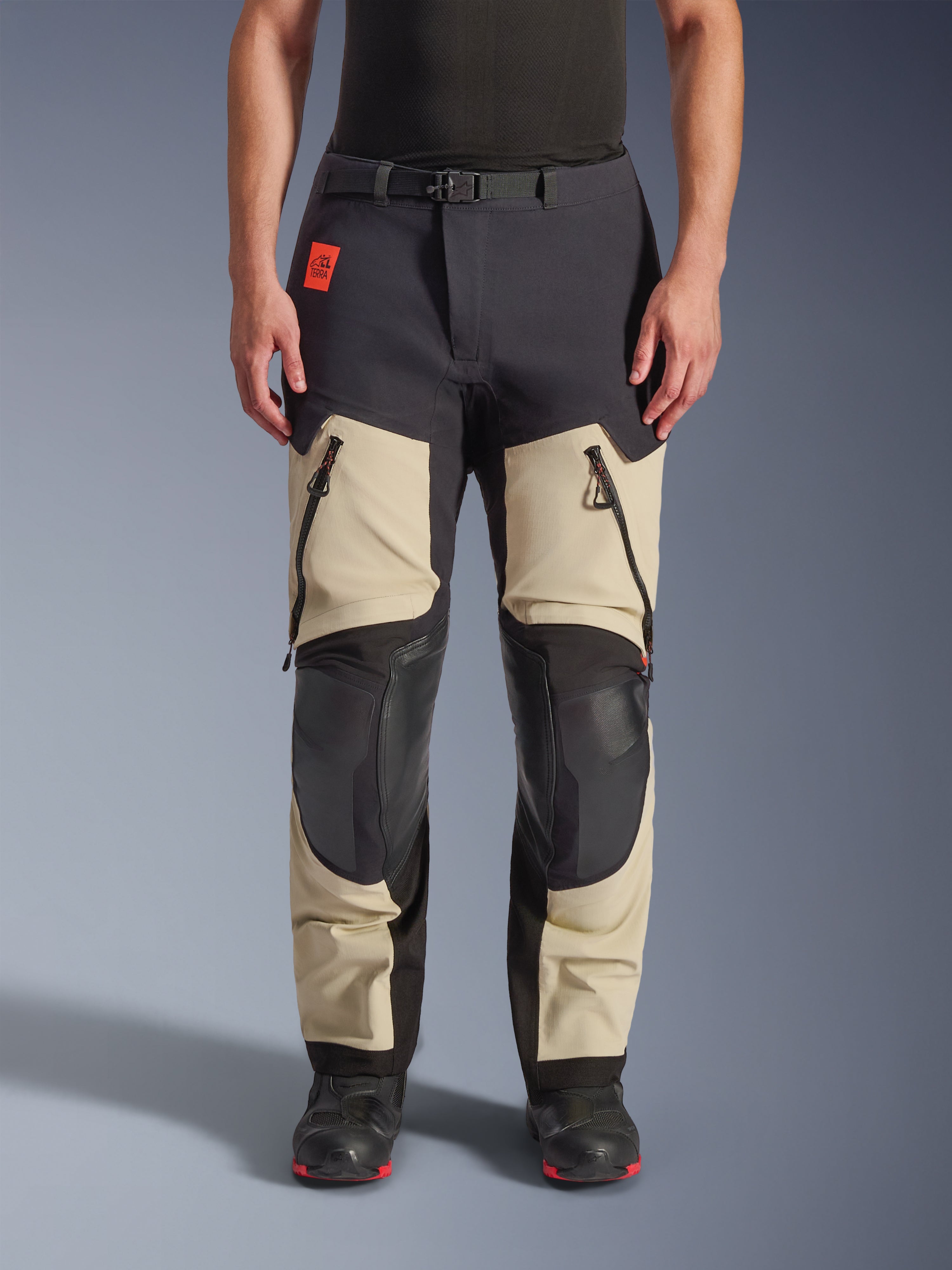 Alpinestars Halo Pro Drystar® XF Laminierte Hose, Adventure-Motorradhose, Mastic Black, Vorderansicht getragen, mit großen Belüftungsöffnungen mit Reißverschluss an den Oberschenkeln, verstärkten schwarzen Kniepaneelen und sandfarbener Textilkonstruktion, kombiniert mit schwarz-roten Motorradstiefeln