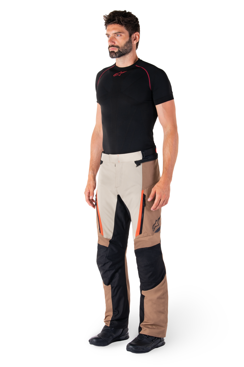 Man wearing Alpinestars ST-1 Waterproof Pants, Adventure Touring Pants, Dark Khaki Sand Black and Red Fluo, styled with a black base layer shirt and black motorcycle boots, featuring reinforced panels and orange zipper accents