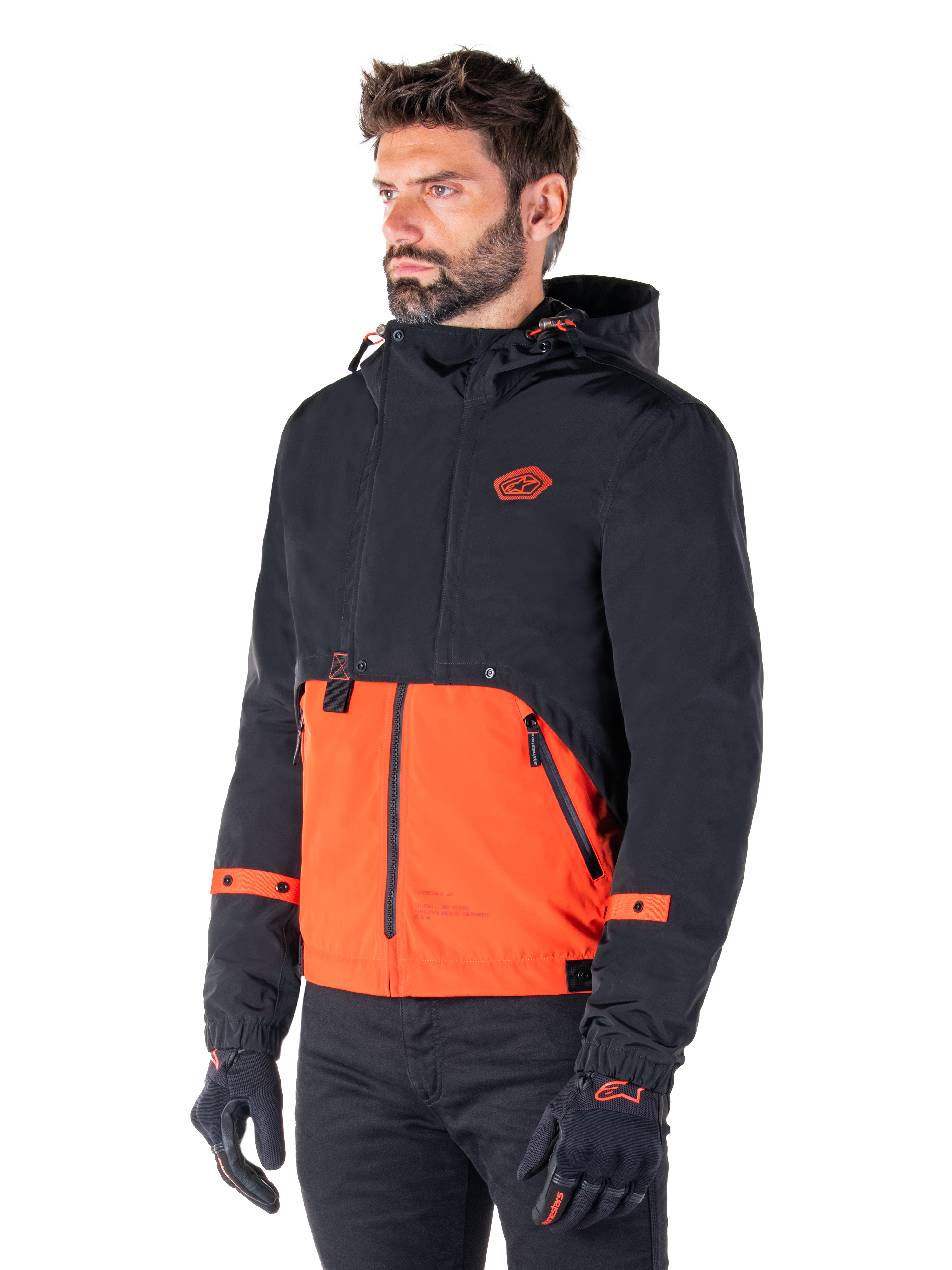 Man wearing the Alpinestars Mootant Waterproof Parka in Spicy Orange and Black, a modular motorcycle jacket with a black hooded upper layer and bright orange lower section, styled with black riding gloves and pants.