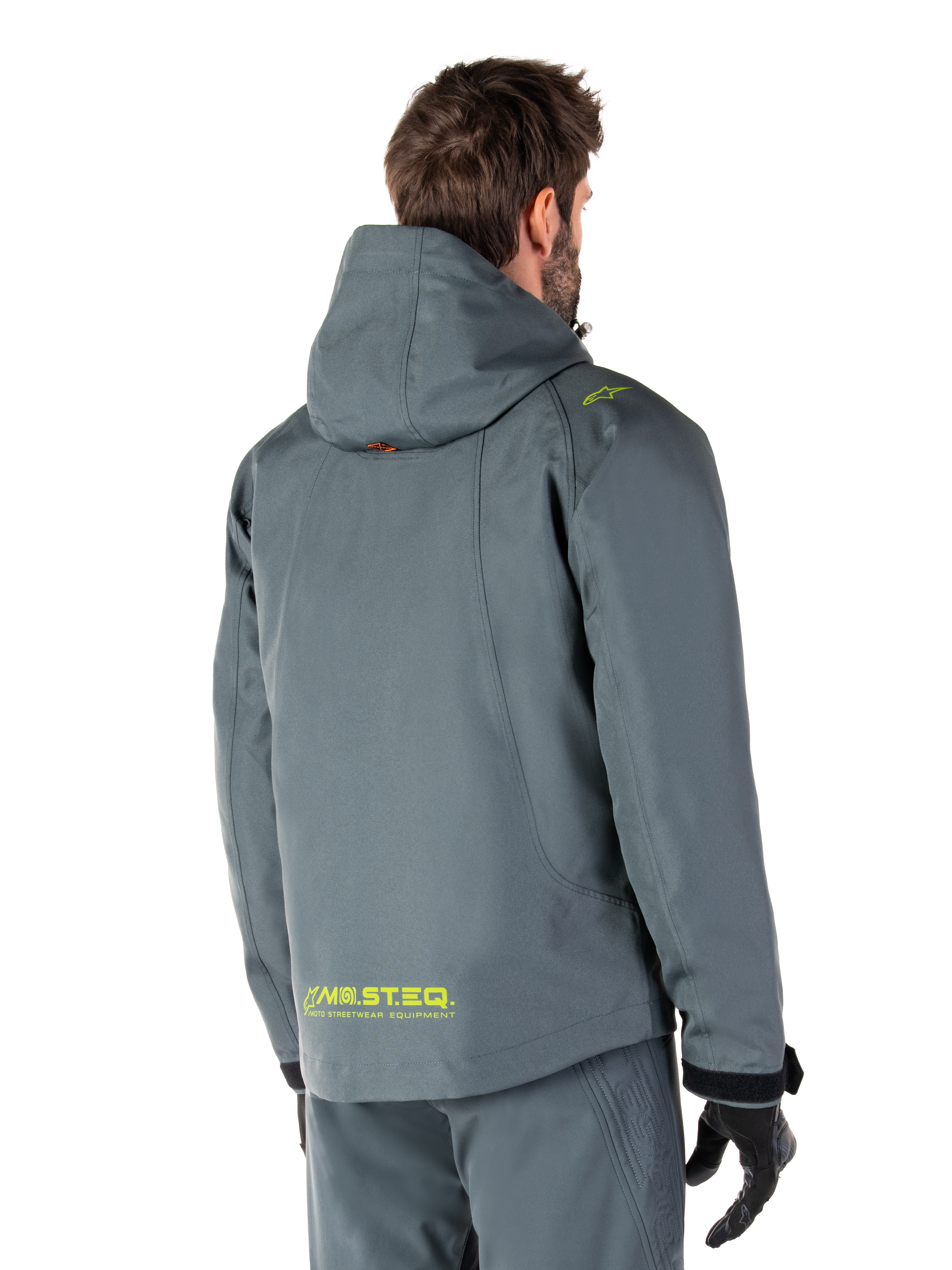 Alpinestars Morush Wasserdichter Parka in Urban Gray, Rückansicht, mit gelbem MO.ST.EQ. Branding am unteren Rücken und an der Schulter, minimalistisches Kapuzendesign, getragen mit passender grauer Motorradhose und schwarzen Handschuhen, urbane Motorrad-Streetwear.