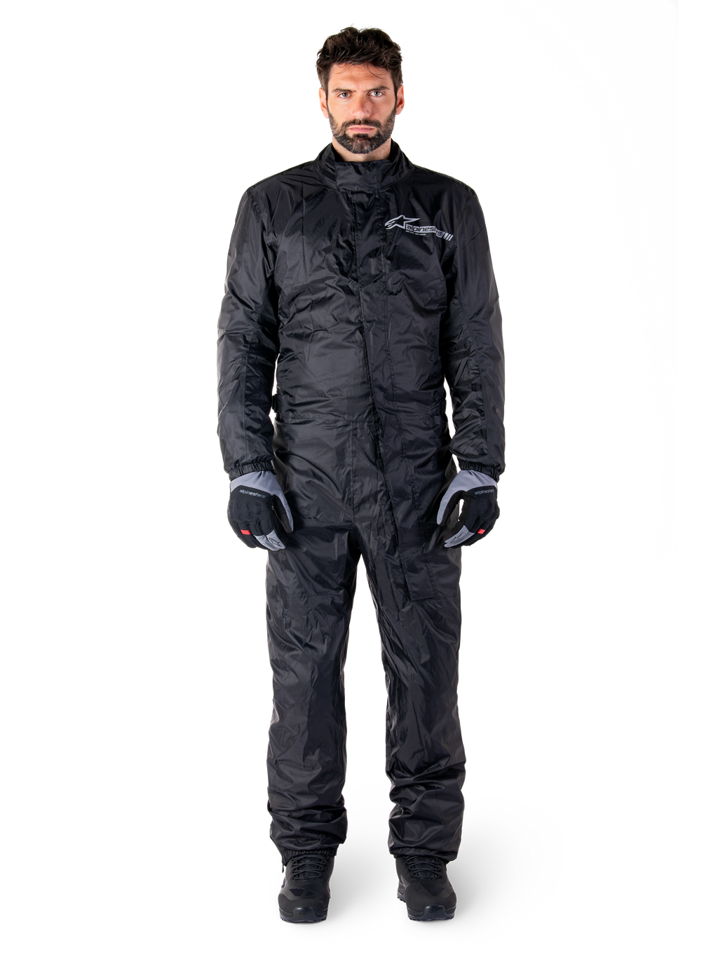 Alpinestars Hurricane Rain V2 Anzug, Ganzkörper-Motorrad-Regenanzug in Schwarz, wasserdichte Schutzausrüstung mit hohem Kragen und Front-Sturmverschluss, weißes Logo auf der Brust, getragen mit schwarz-grauen Handschuhen und Stiefeln