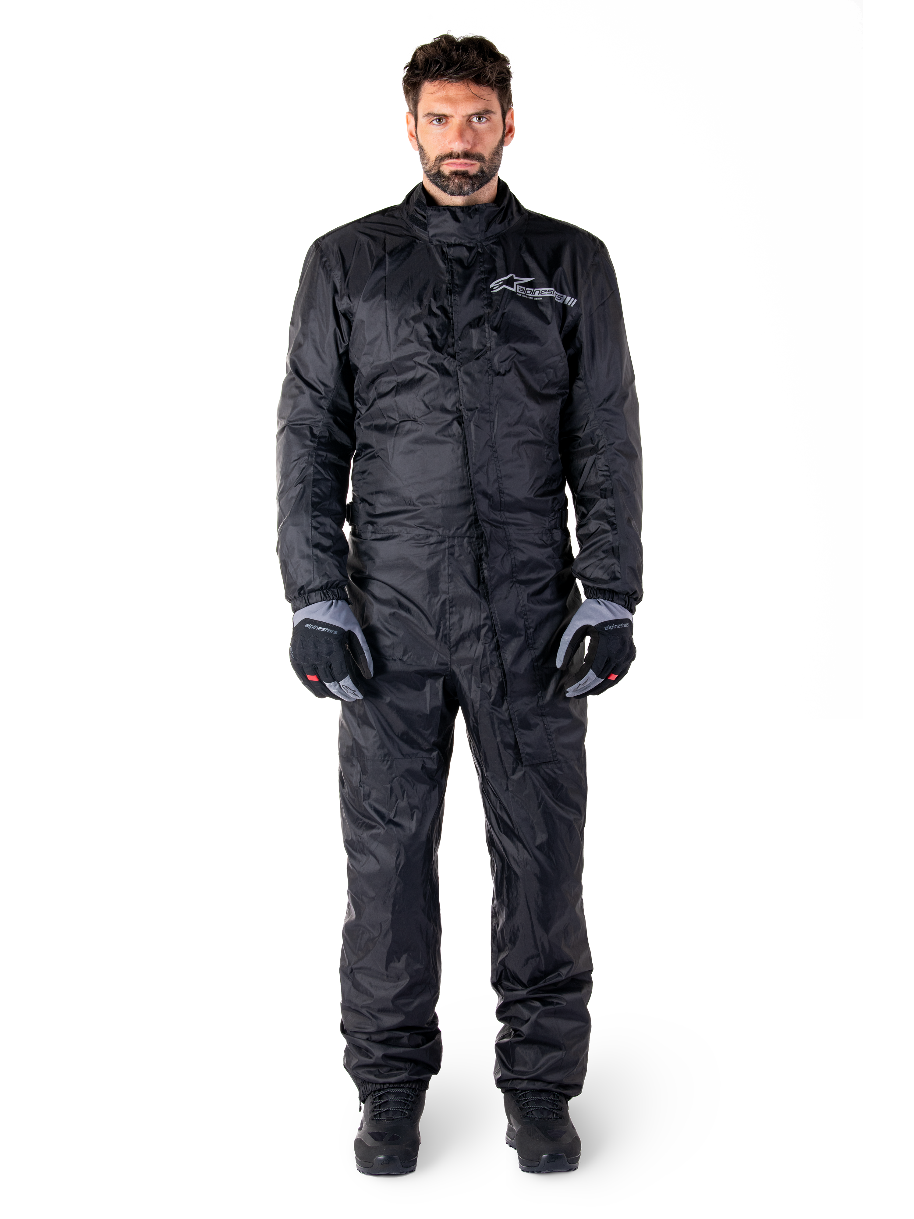 Alpinestars Hurricane Rain V2 Anzug, Ganzkörper-Motorrad-Regenanzug in Schwarz, wasserdichte Schutzausrüstung mit hohem Kragen und Front-Sturmverschluss, weißes Logo auf der Brust, getragen mit schwarz-grauen Handschuhen und Stiefeln