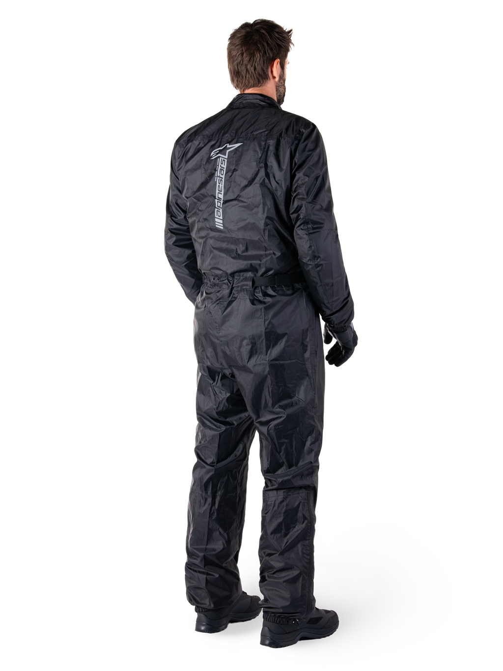 Alpinestars Hurricane Rain V2 Suit, schwarzer einteiliger wasserdichter Motorrad-Regenanzug, Rückansicht mit großem vertikalem reflektierendem Logo, leichtes windabweisendes Gewebe, getragen mit schwarzen Handschuhen und Stiefeln