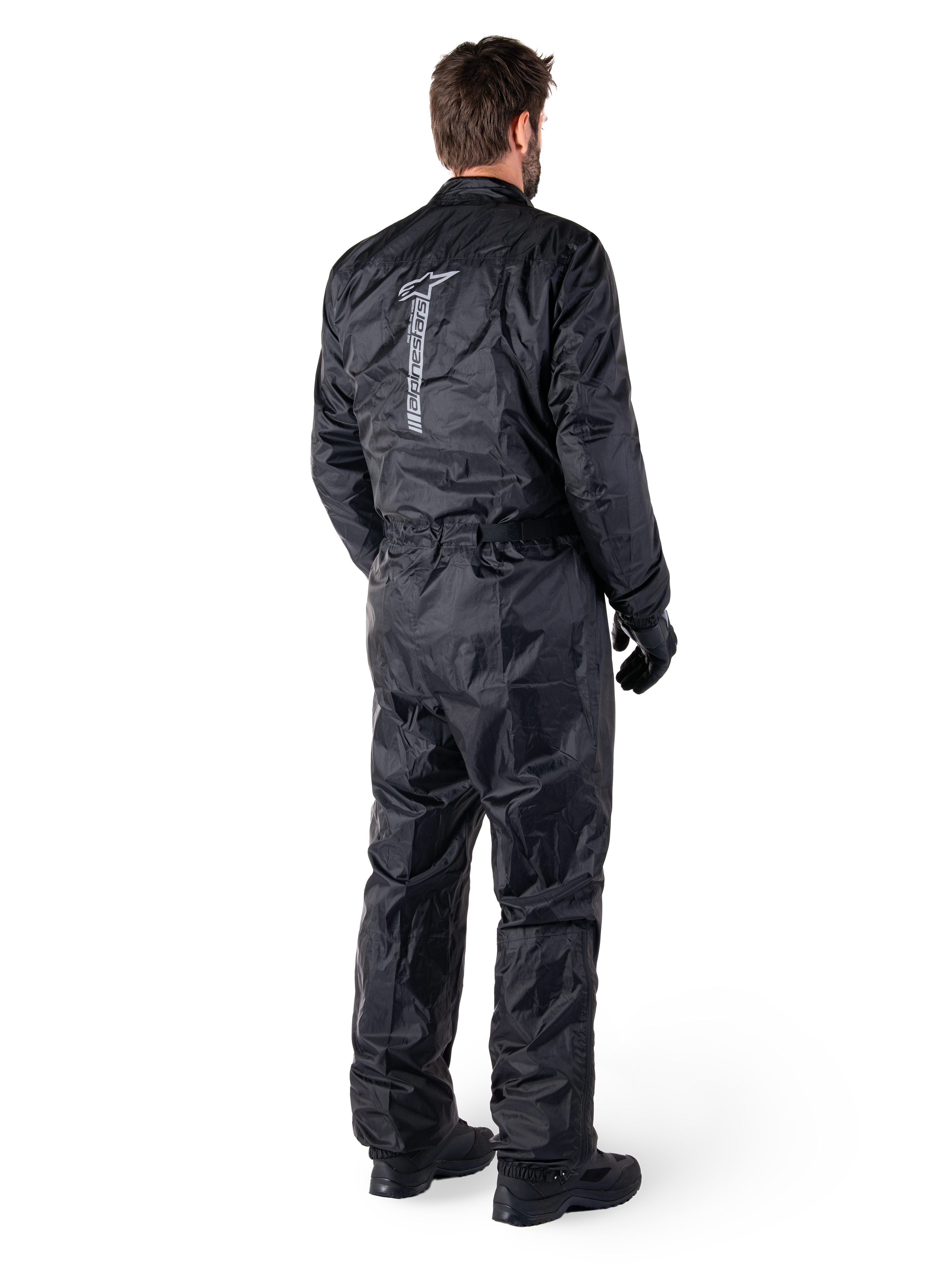Alpinestars Hurricane Rain V2 Suit, schwarzer einteiliger wasserdichter Motorrad-Regenanzug, Rückansicht mit großem vertikalem reflektierendem Logo, leichtes windabweisendes Gewebe, getragen mit schwarzen Handschuhen und Stiefeln