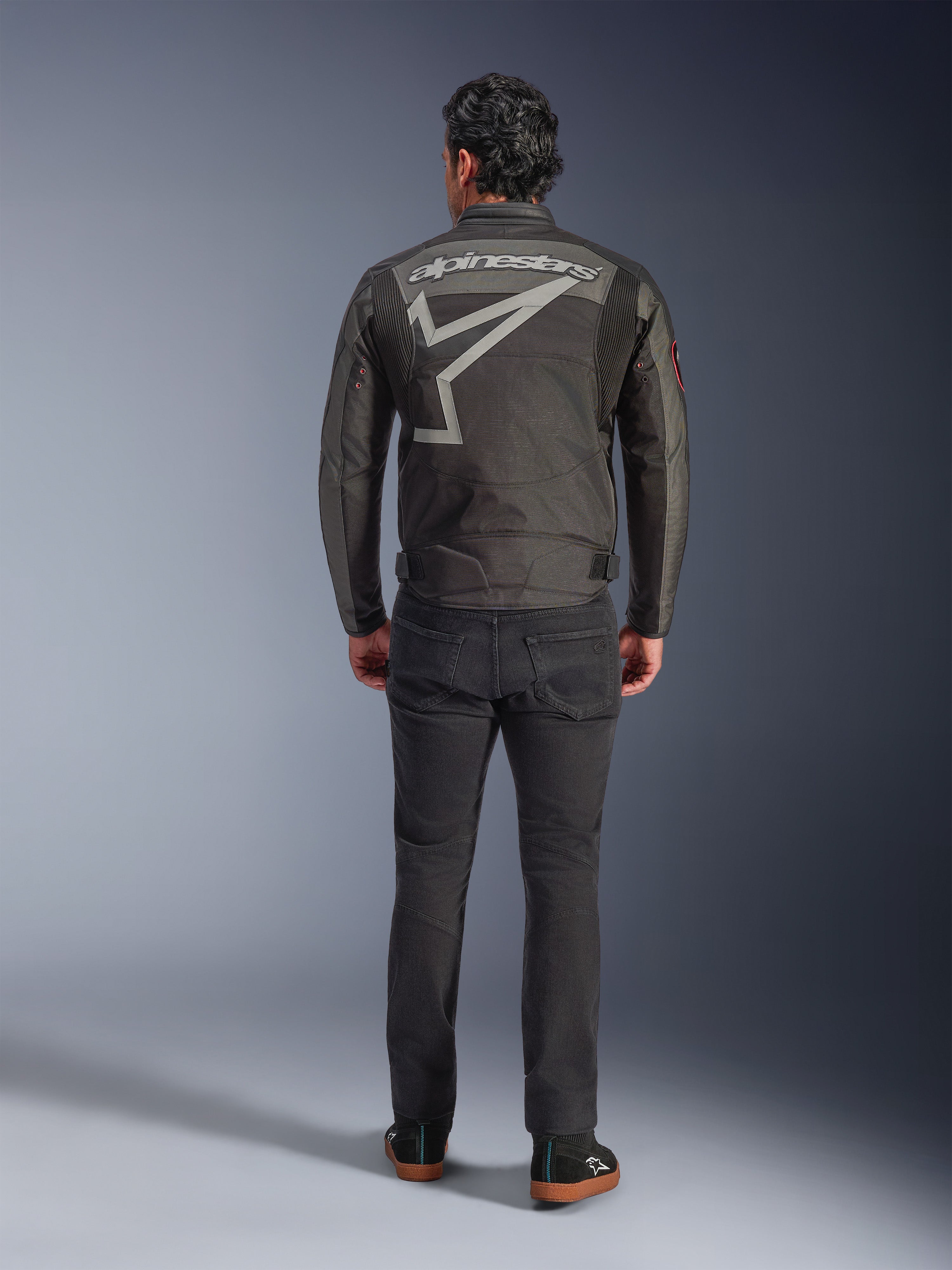 Alpinestars Axton Jacke, Motorradjacke, Schwarz Anthrazit Rot Tangerine, Rückansicht mit großem grauem Alpinestars-Logo und Sterngrafik, Akkordeon-Schulterpaneele, getragen mit schwarzer Hose und schwarzen Freizeitschuhen.