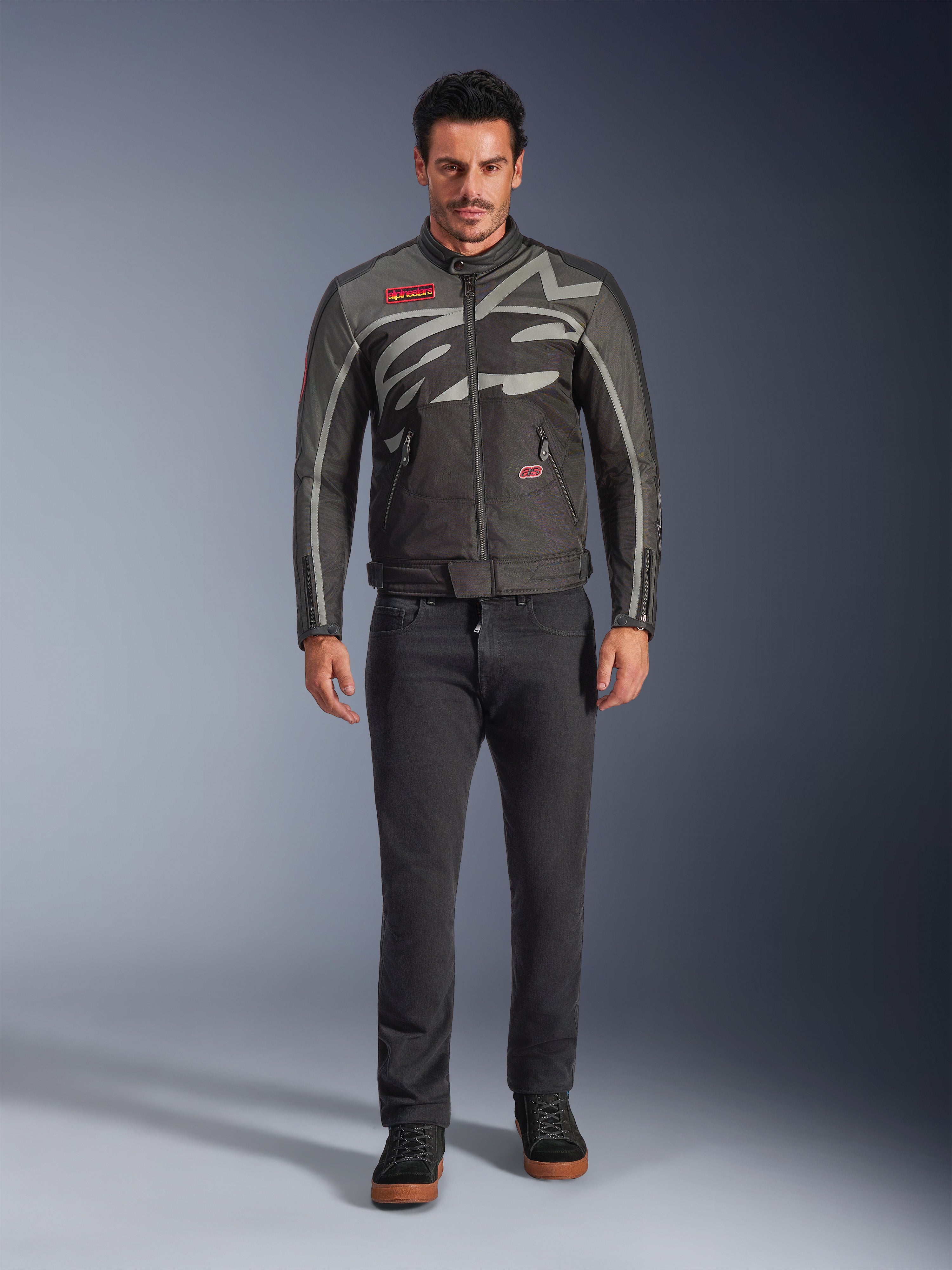Alpinestars Axton Jacke, Motorradjacke, Schwarz Anthrazit Rot Tangerine, getragen von einem Model mit dunkler Hose, mit umlaufendem grauen Alpinestars-Logo, roten Markenaufnähern und wasserabweisender Textil-Außenschicht für urbane Straßenfahrten