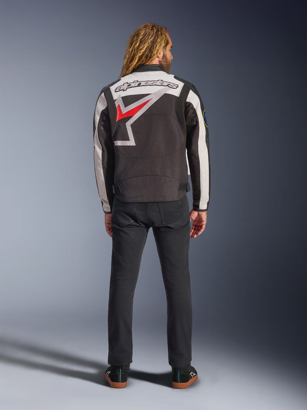 Alpinestars Axton Jacke, Motorradjacke, Schwarz, Off-White und Bright Red, Rückansicht an einem Model, mit einem großen Alpinestars-Sternlogo auf dem Rücken, Off-White-Ärmeleinsätzen, getragen mit einer schwarzen Hose, von Vintage-Rennsportbekleidung inspiriertes Design