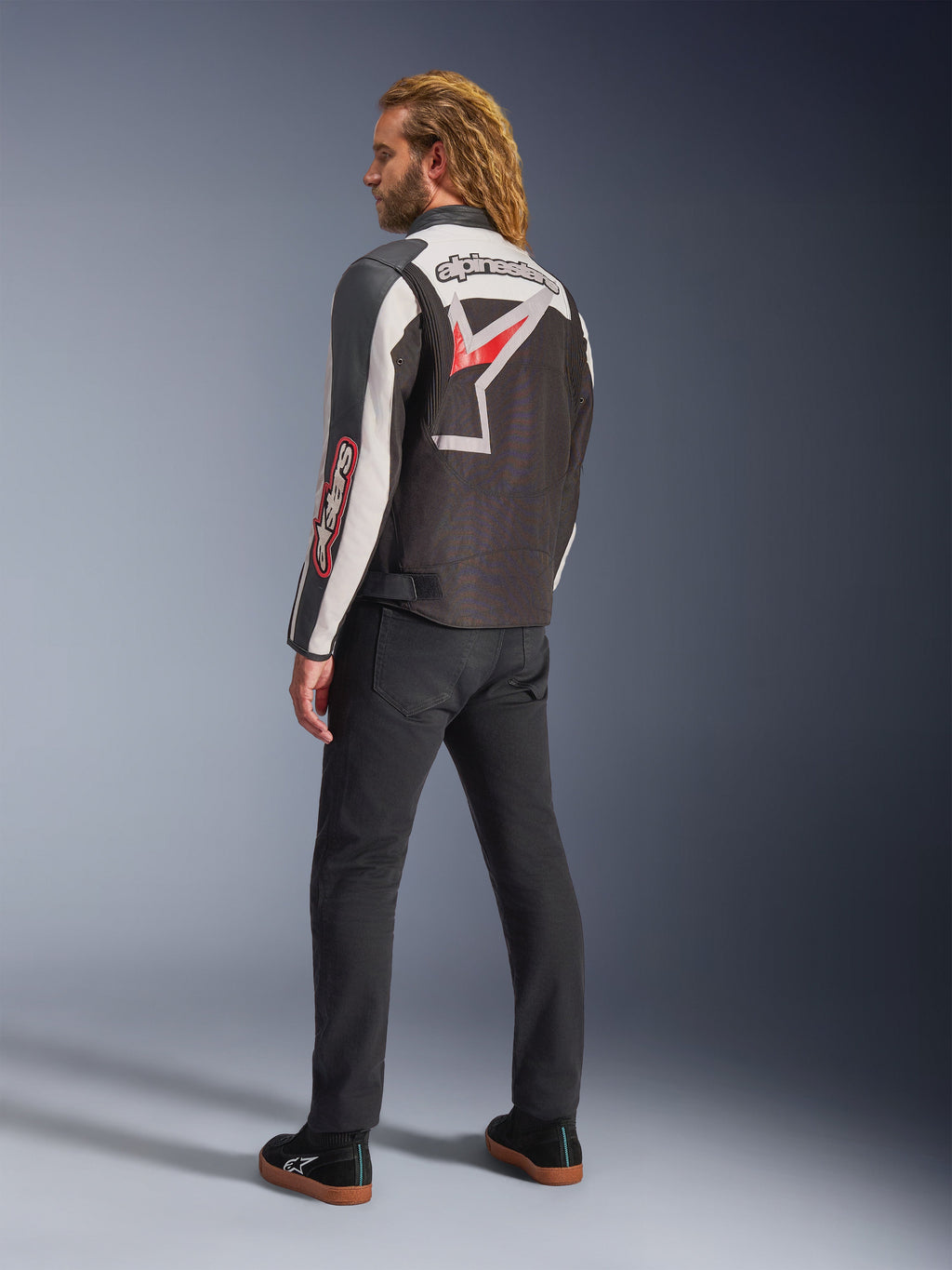 Alpinestars Axton Jacke, Motorradjacke, Schwarz, Off-White und leuchtendes Rot, Rückansicht getragen von einem männlichen Model, mit großem Alpinestars Stern-Logo und Schriftzug auf dem Rücken, Vintage-Rennsport-inspiriertes Design mit Akkordeon-Einsätzen, kombiniert mit schwarzer Hose und Sneakern