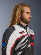 Mann mit langen Haaren und Bart trägt die Alpinestars Axton Jacke, Motorradjacke, Schwarz, Off-White und Bright Red, vom Vintage-Rennsport inspiriertes Design mit Marken-Patches und umlaufendem Logo, Nahaufnahme in der Profilansicht, urbaner zeitgenössischer Stil