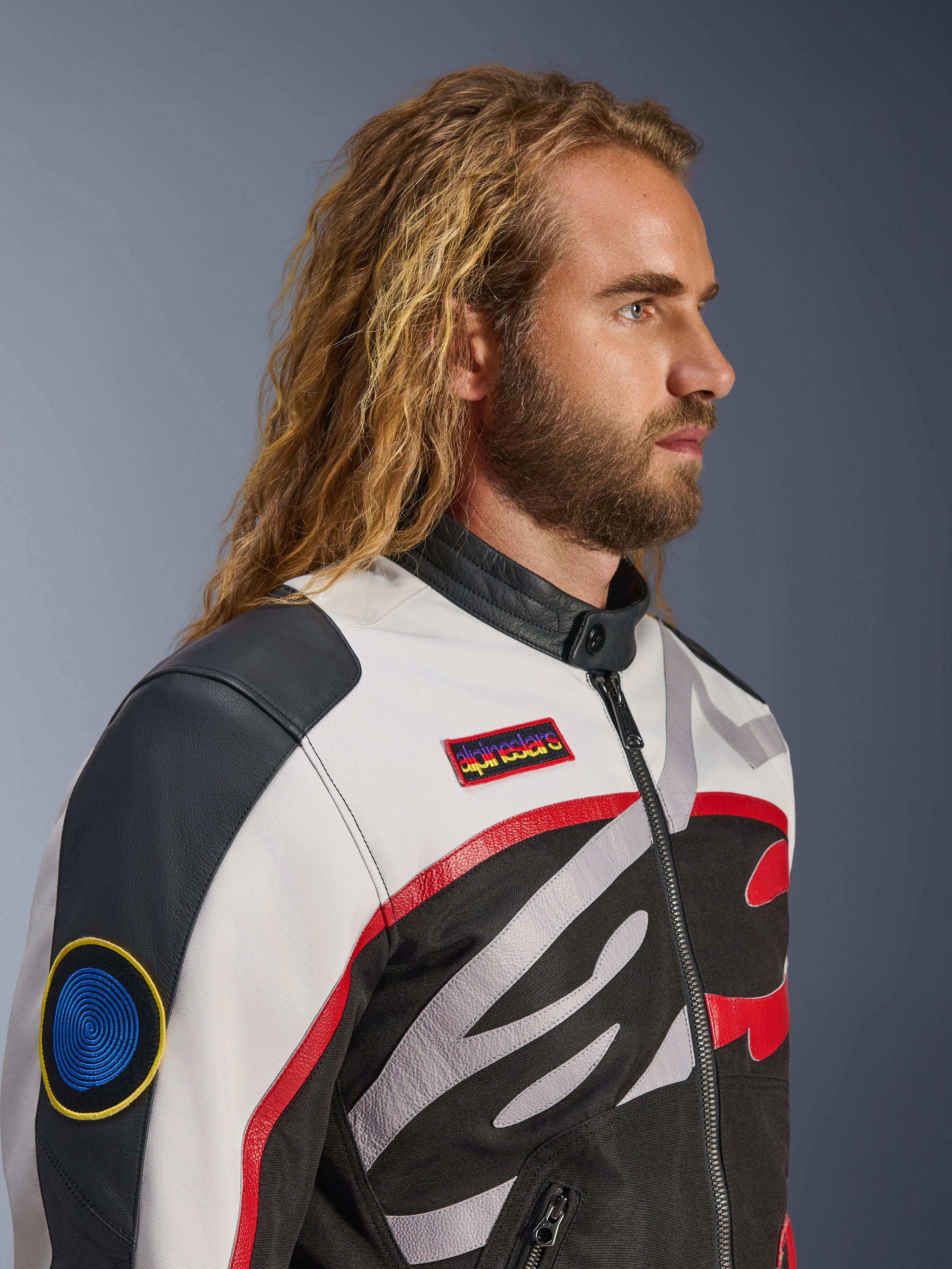 Mann mit langen Haaren und Bart trägt die Alpinestars Axton Jacke, Motorradjacke, Schwarz, Off-White und Bright Red, vom Vintage-Rennsport inspiriertes Design mit Marken-Patches und umlaufendem Logo, Nahaufnahme in der Profilansicht, urbaner zeitgenössischer Stil