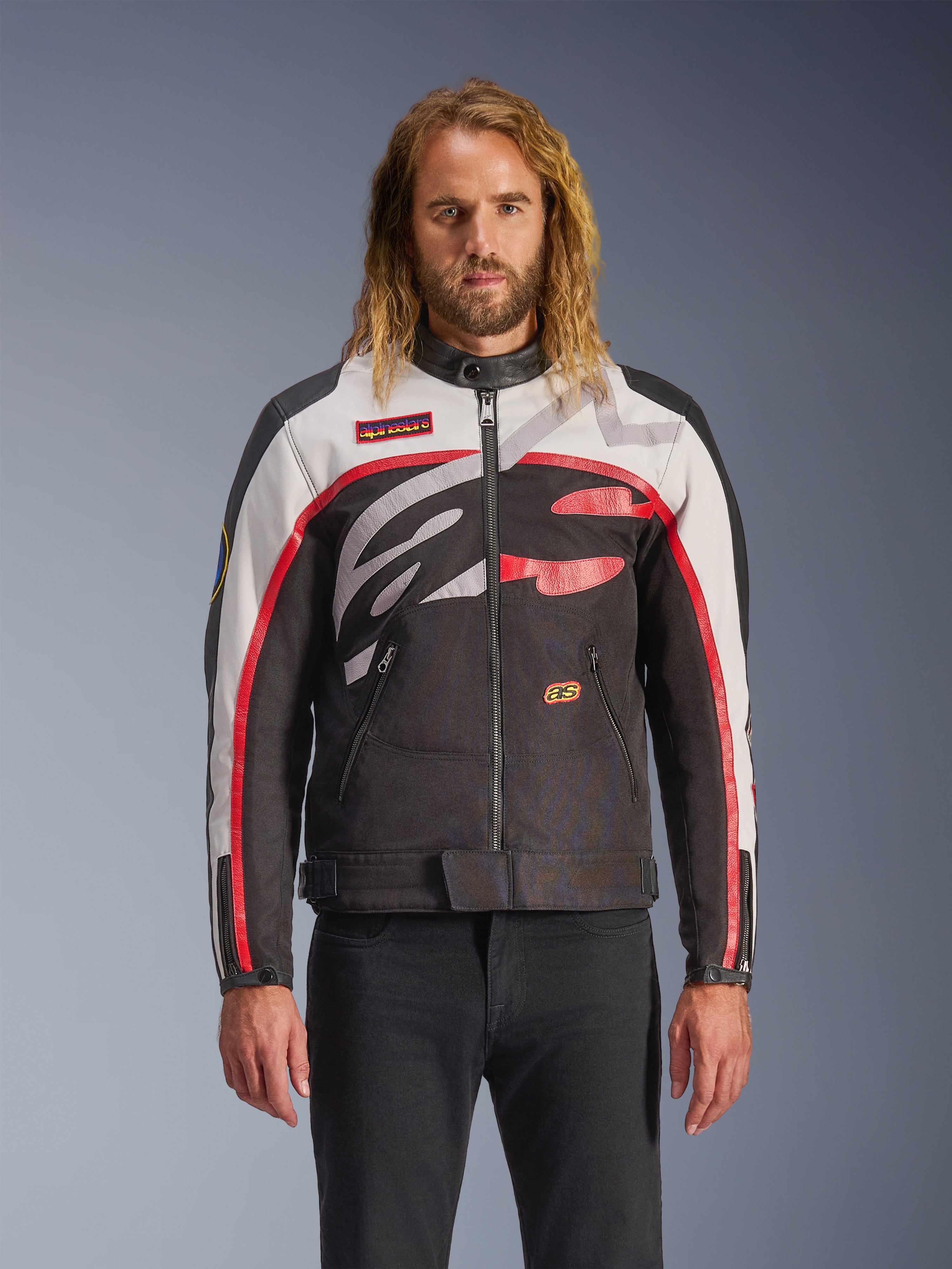 Alpinestars Axton Jacke, Motorradjacke, Schwarz, Off-White und leuchtendes Rot, mit markanten abstrakten Mustern auf der Brust und umlaufendem Logo, rot gestreifte Ärmel, Vintage-inspiriertes Design mit minimalistischem Kragen, getragen von einem Model mit schwarzen Jeans.