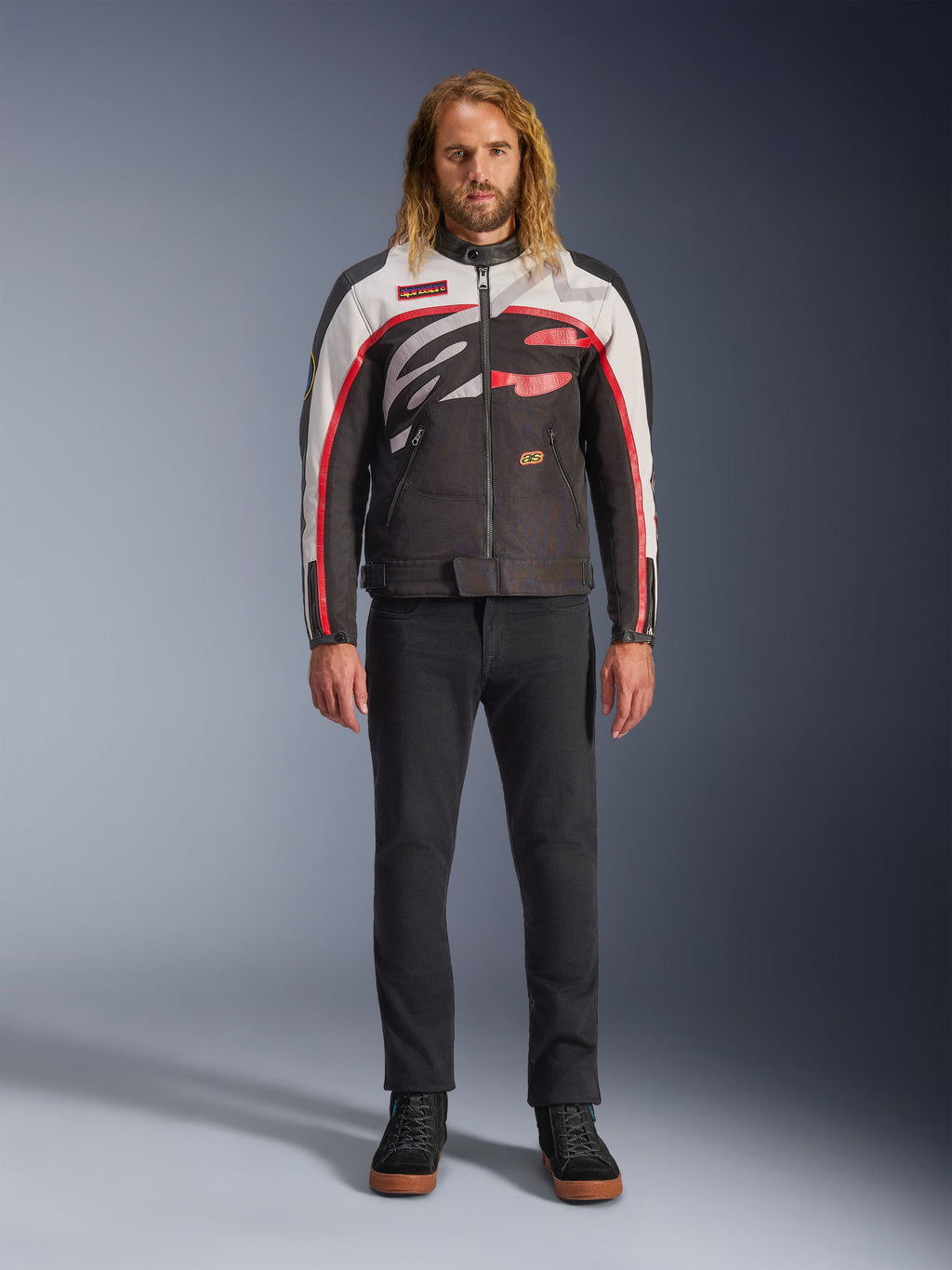 Alpinestars Axton Jacke, Motorradjacke, Schwarz, Off-White und Leuchtendes Rot, getragen von einem Model mit schwarzer Hose und Sneakern, mit einem markanten, umlaufenden Logo und einem vom Vintage-Rennsport inspirierten Design