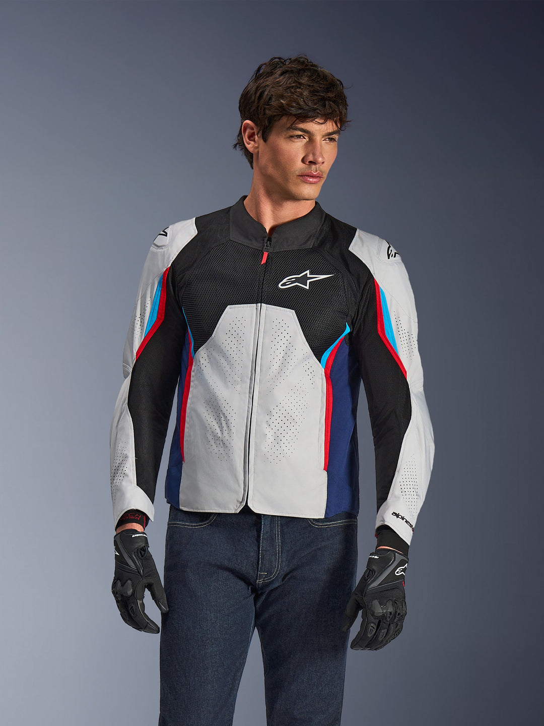 Alpinestars AST V3 Air Jacke in Silber Deep Blue Bright Red, getragen von einem Model mit dunkler Jeans und schwarzen Handschuhen, mit perforierten silbernen Panels, Belüftung aus schwarzem Mesh und sportlichen roten und blauen Akzenten.