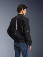 Alpinestars Flight Air Jacke, schwarze Motorradjacke, Rückansicht mit großem vertikalem weißem Logo und Mesh-Belüftungseinsätzen, Bomber-Stil mit geripptem Saum, getragen mit blauen Jeans, urbane zeitgenössische Motorradbekleidung