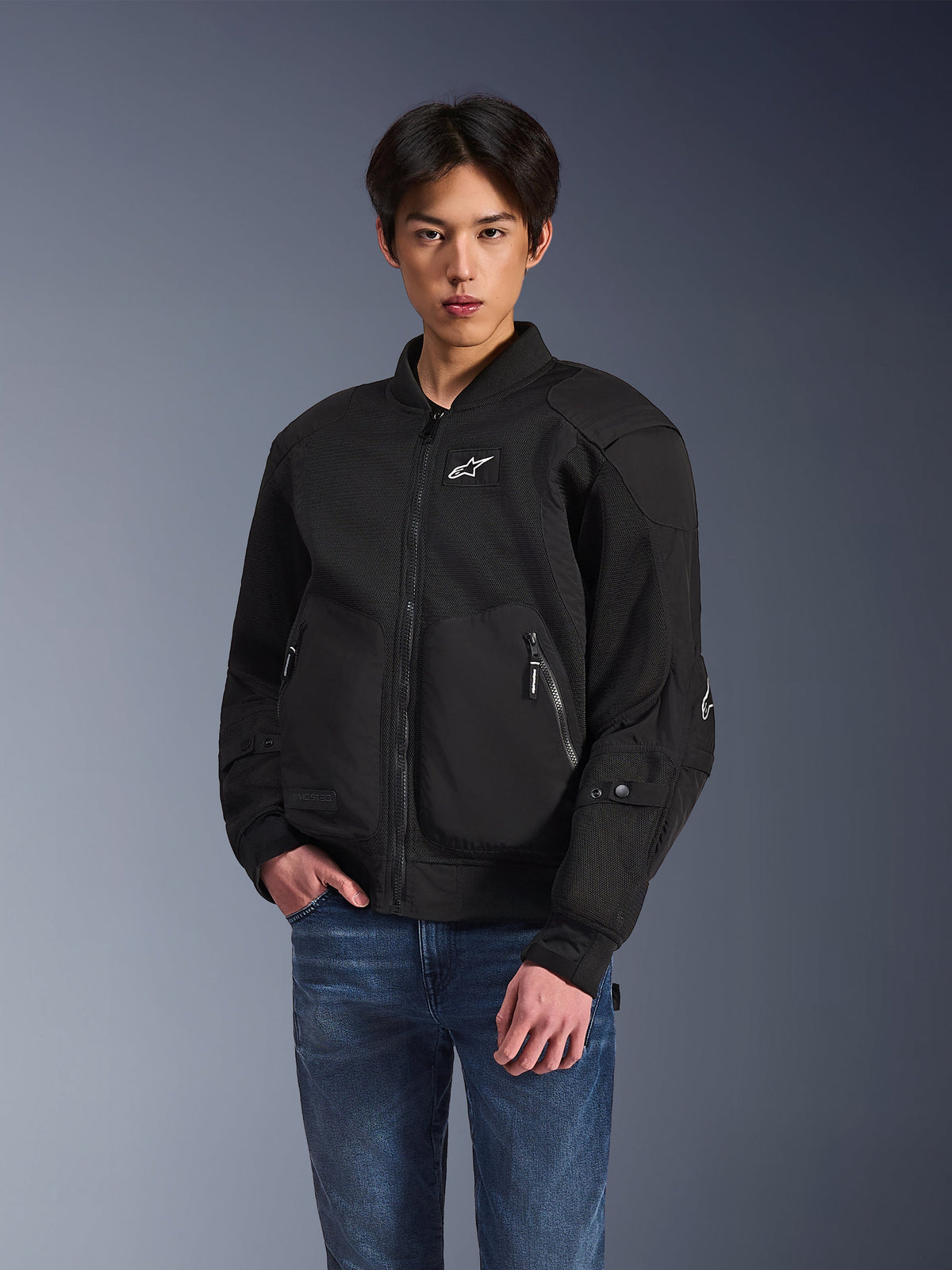 Alpinestars Flight Air Jacke, schwarze Urban-Motorradjacke, getragen von einem Mann mit blauen Jeans, im Bomberjacken-Stil mit Mesh-Einsätzen, Frontreißverschluss und weißem Logo auf der Brust, entwickelt für Stadtfahrten bei heißem Wetter