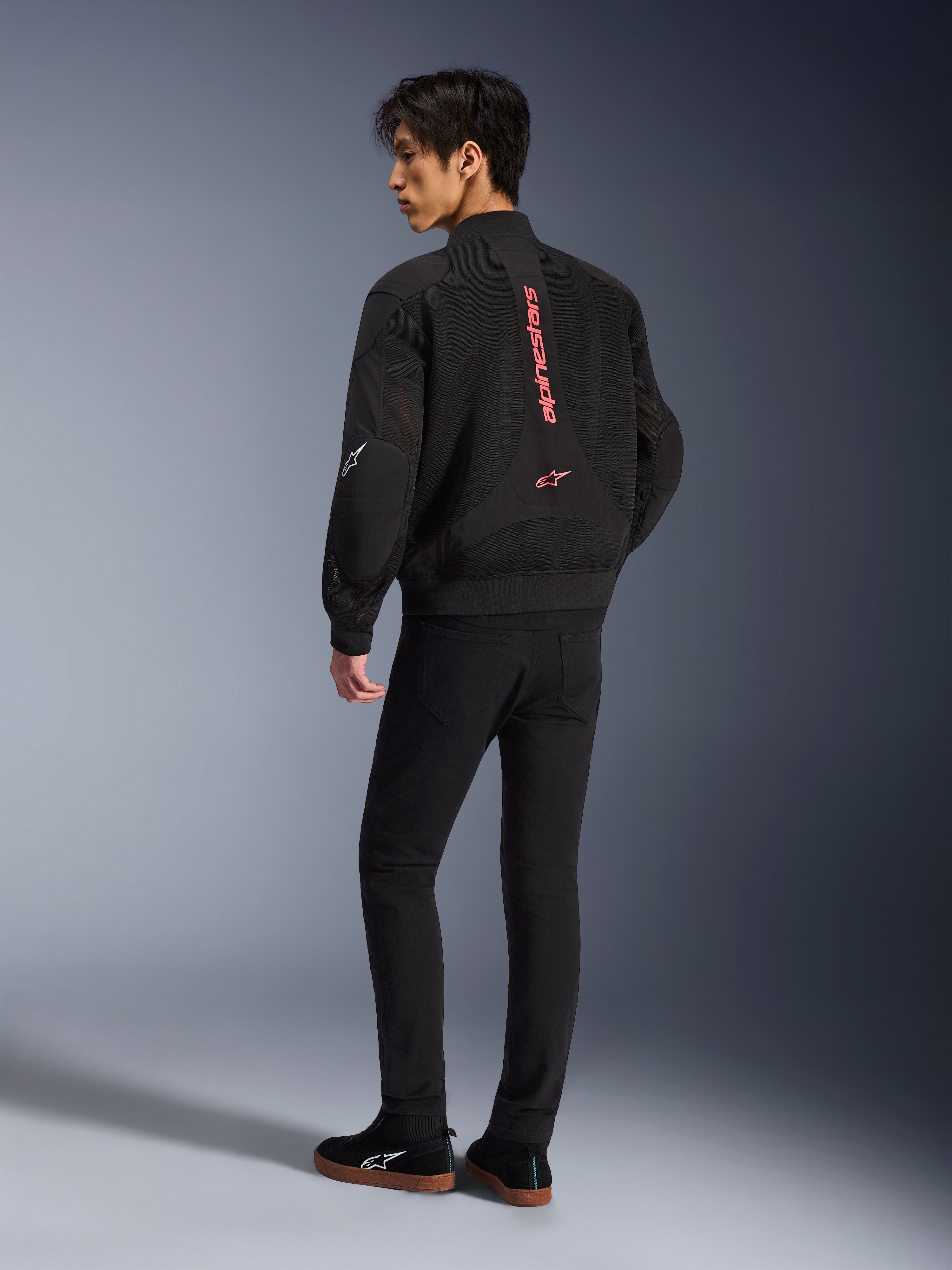 Männliches Model trägt die Alpinestars Flight Air Jacke in Black Bright Red, Rückansicht mit vertikalem rotem Branding auf dem Rücken und 3D-Mesh-Konstruktion, kombiniert mit schwarzer Hose und Sneakern, urbaner Motorrad-Stil