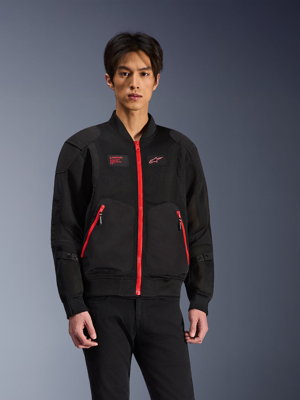 Alpinestars Flight Air Jacke in Schwarz und leuchtendem Rot, urbane Motorradjacke im Bomber-Stil, getragen von einem Model, mit Mesh-Belüftungseinsätzen, einem auffälligen roten Frontreißverschluss, roten Taschendetails und Branding auf der Brust, kombiniert mit einer schwarzen Hose