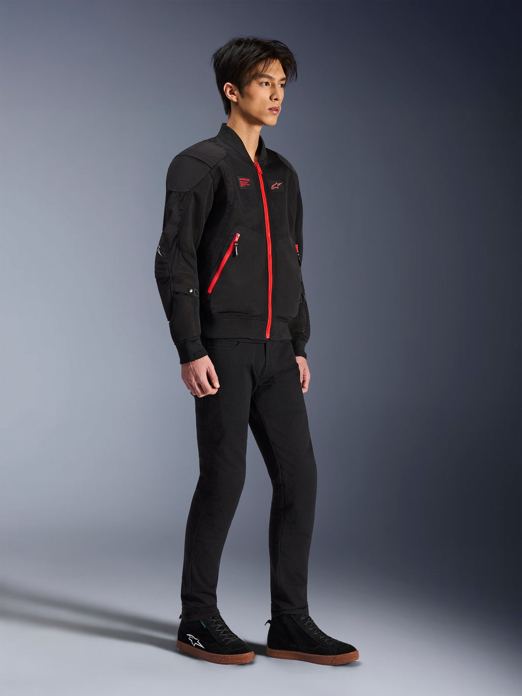 Alpinestars Flight Air Jacke, Schwarz und leuchtendes Rot, urbane Motorradjacke im Bomber-Stil, getragen von einem Modell, mit 3D-Mesh-Paneelen und kontrastierenden roten Reißverschlüssen, kombiniert mit schwarzer Hose und lässigen Motorradschuhen, moderne Streetwear-inspirierte Schutzausrüstung