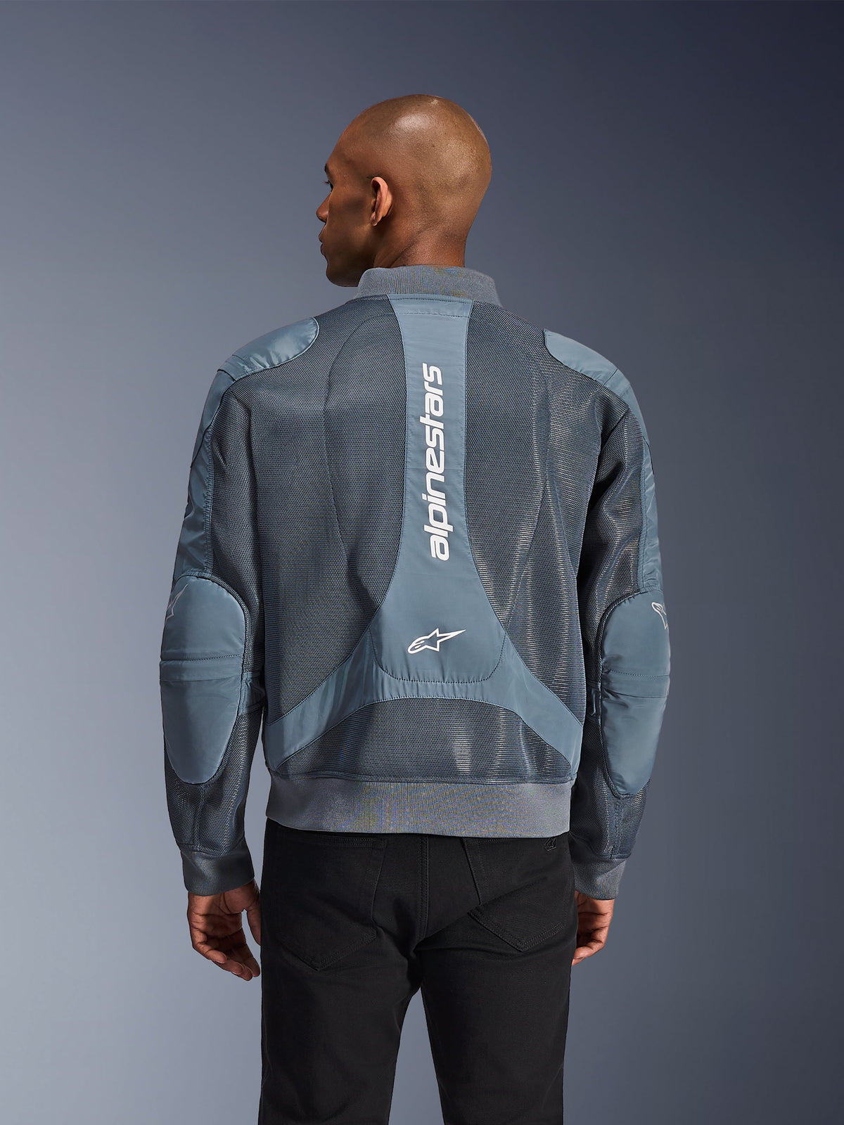Alpinestars Flight Air Jacke in Storm Gray, Rückansicht an einem Model, urbane Motorradjacke mit erweiterten 3D-Mesh-Einsätzen, vertikalem Alpinestars-Branding auf dem Rücken sowie geripptem Saum und Kragen im Bomber-Stil.