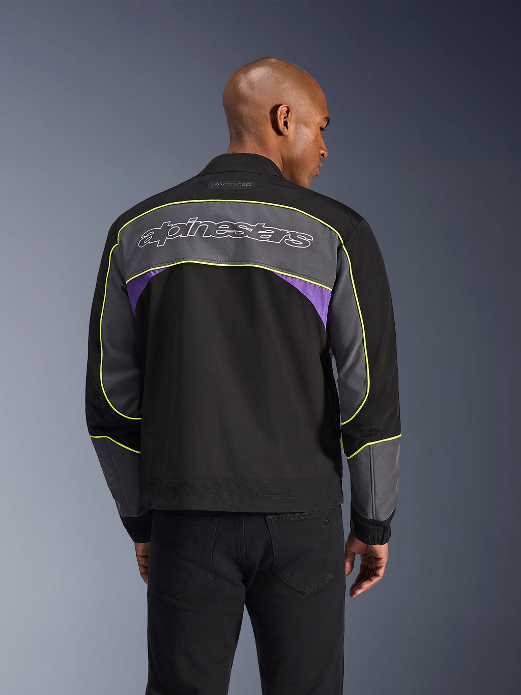 Alpinestars Unite Jacke, Urbane Motorradjacke, Schwarz Asphalt Violett, Rückansicht mit grauen Panels und violetten Akzenten, limettengelbe Paspelierung, großer Alpinestars-Schriftzug auf dem oberen Rücken, minimalistisches Streetwear-Design