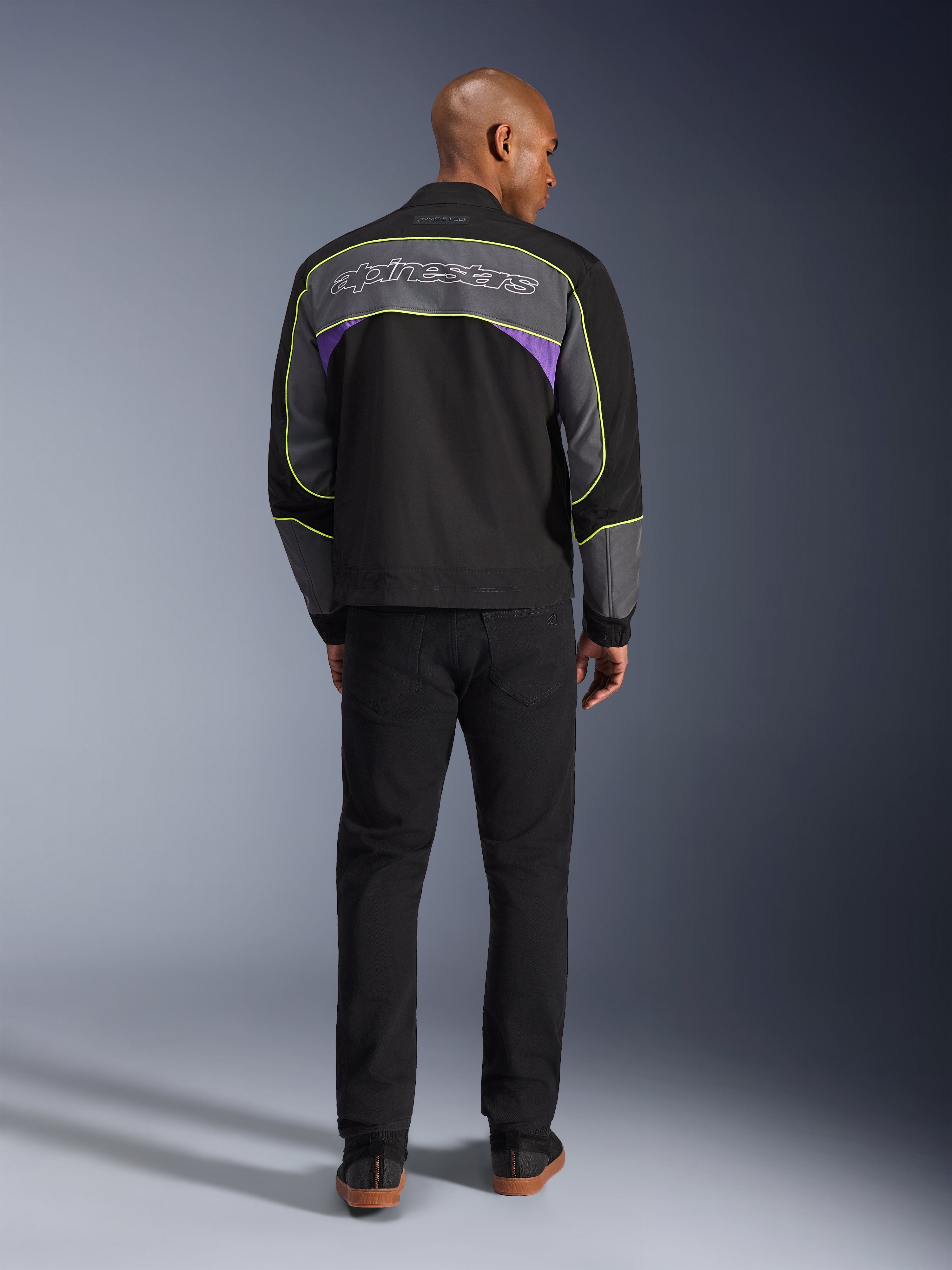 Alpinestars Unite Jacke, urbane Motorradjacke in Black Asphalt Violet, Rückansicht mit grauen Rückenbahnen, lila Akzenten und neongelben Paspeln, Alpinestars-Schriftzug über den Schultern, getragen mit schwarzer Hose