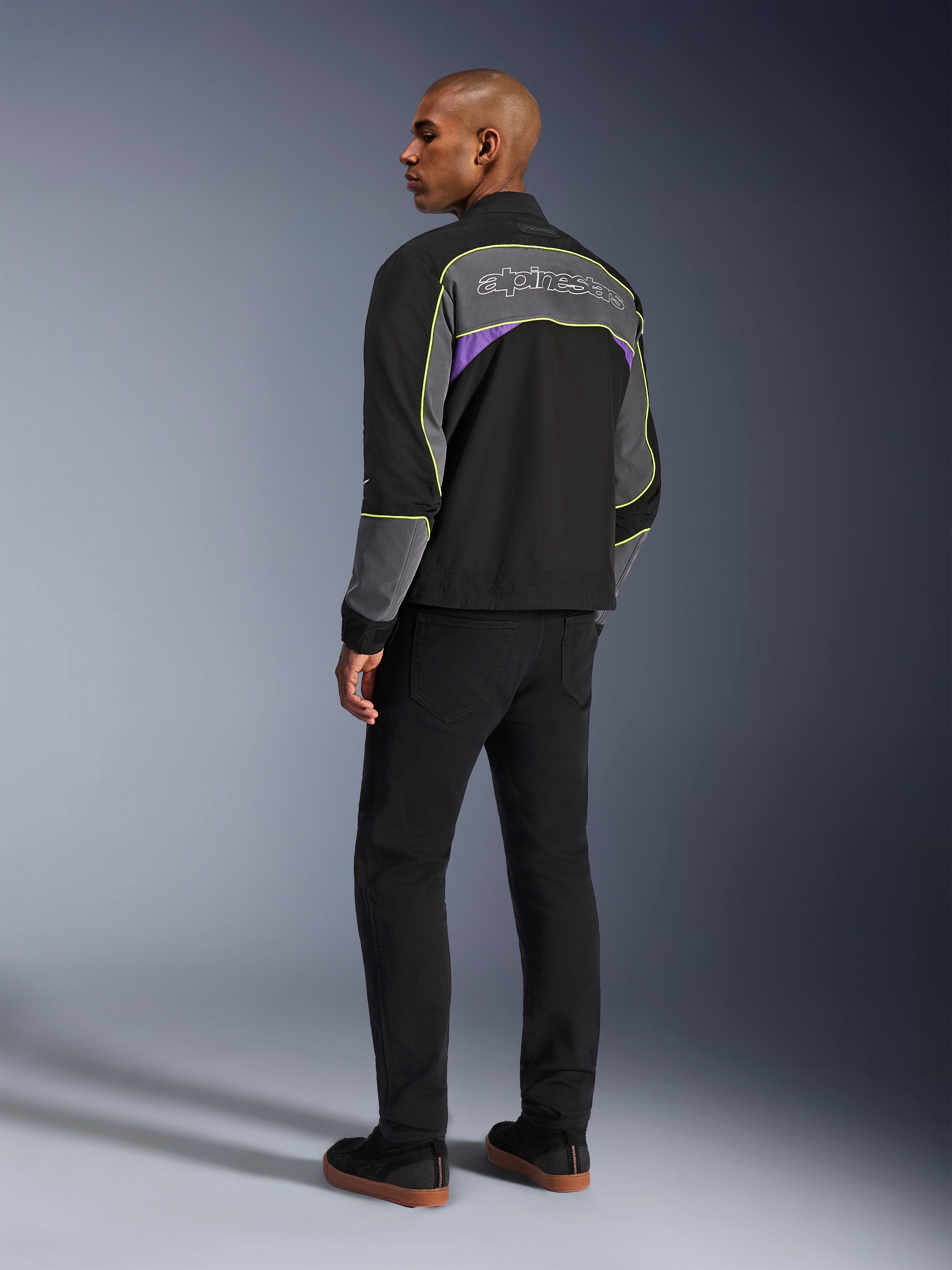 Alpinestars Unite Jacke, urbane Motorradjacke, Black Asphalt Violet, Dreiviertel-Rückansicht am Modell, schwarze Softshell mit grauen Einsätzen und violetten Akzenten, neongelbe reflektierende Paspelierung, Alpinestars-Logo auf dem oberen Rücken, kombiniert mit schwarzer Hose und schwarzen Freizeitschuhen