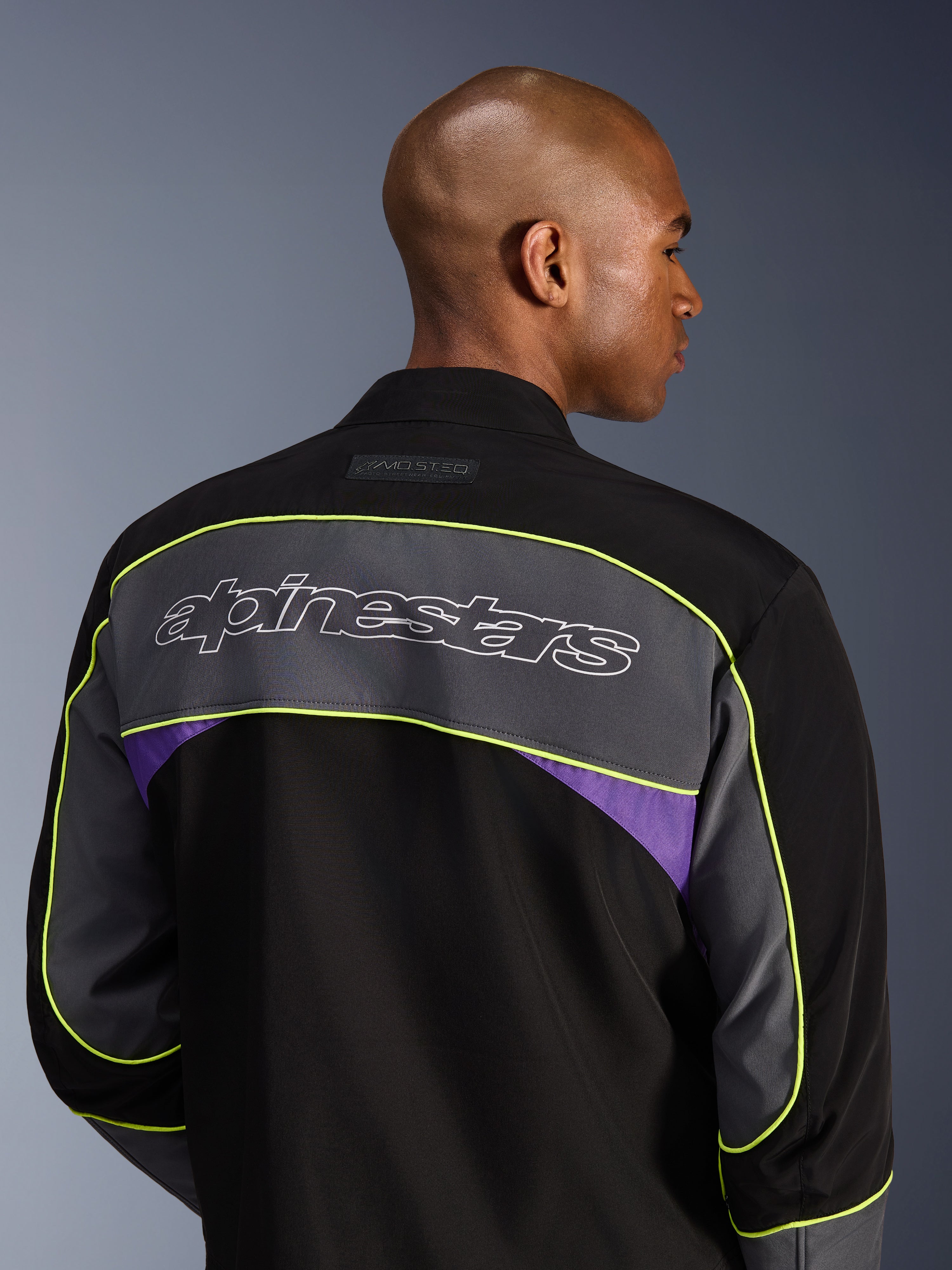 Alpinestars Unite Jacke, urbane Motorradjacke, Black Asphalt Violet, Rückansicht mit einem großen grauen Schulterpanel mit Alpinestars-Logo, neongelben Paspeln und violetten Akzenten auf einer 3-lagigen Softshell-Konstruktion