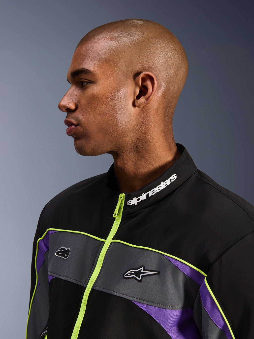 Alpinestars Unite Jacke, Black Asphalt Violet, Profilansicht eines männlichen Modells mit Fokus auf den hohen Kragen mit weißem Alpinestars-Branding, neongelber Reißverschluss, graue Brustpaneele mit Logos, violette Akzente, minimalistisches urbanes Motorrad-Streetwear-Styling