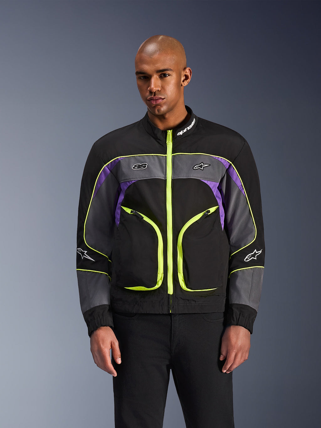 Alpinestars Unite Jacke, urbane Motorradjacke, Schwarz, Asphalt und Violett mit neongelben Akzenten, große Fronttaschen mit neongelben Reißverschlüssen, hoher Kragen mit weißem Logo, getragen von einem Modell, moderne Schutzausrüstung im Streetwear-Stil