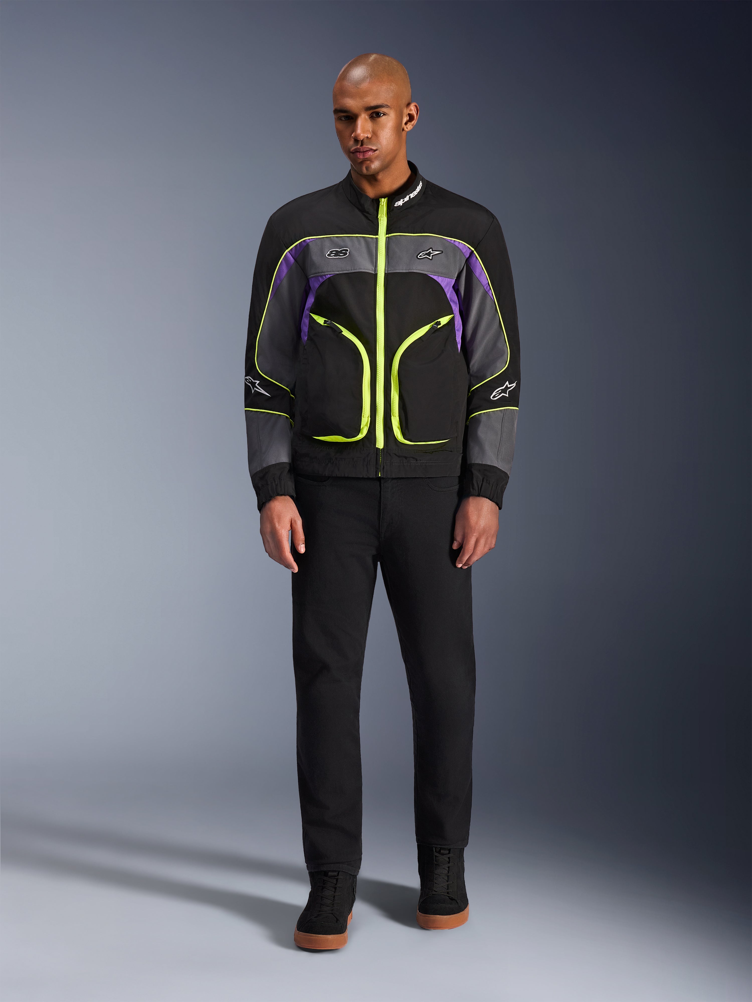 Männliches Model trägt die Alpinestars Unite Jacke in Black Asphalt Violet, mit schwarzer Basis, grauen und lila Einsätzen sowie leuchtend neongelben Reißverschlüssen und Details, kombiniert mit schwarzer Hose und Schuhen, minimalistische urbane Motorradjacke.