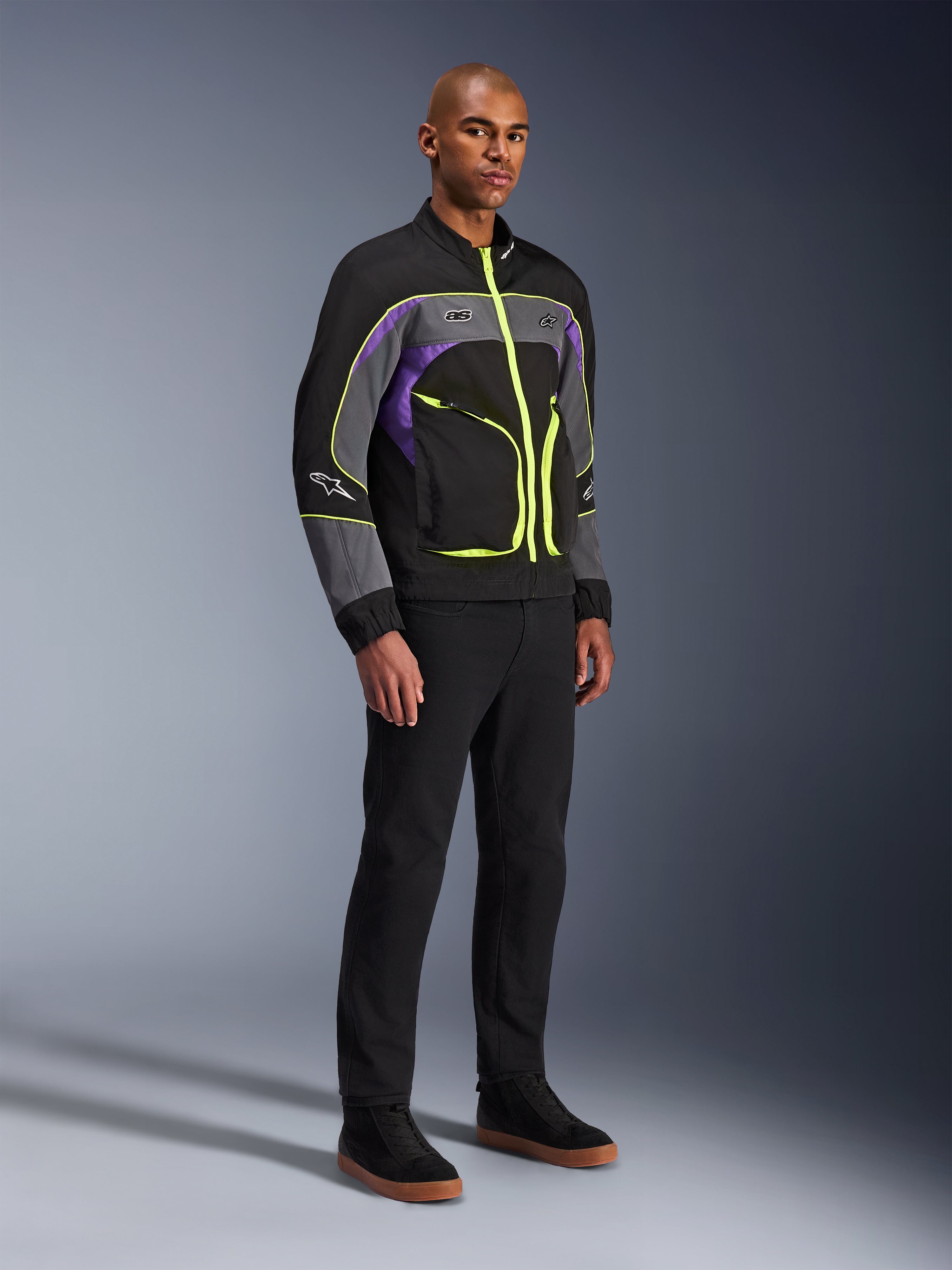 Alpinestars Unite Jacke, urbane Motorradjacke, Black Asphalt Violet mit neongelben Akzenten, getragen von einem Model mit schwarzer Hose und Sneakern, zeitgenössischer Streetwear-Stil für urbane Fahrten