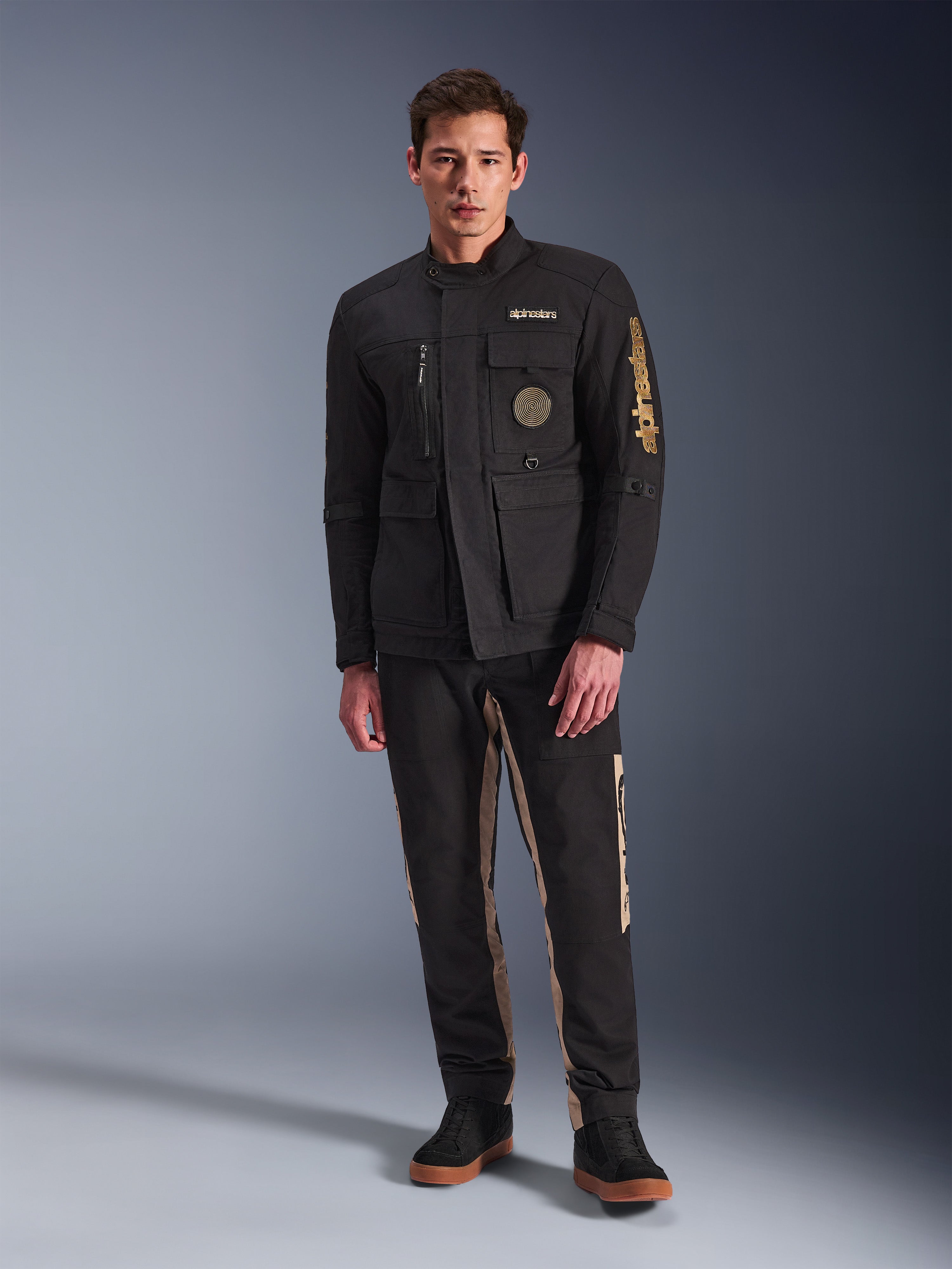 Male model wearing the Alpinestars Dice Jacket, Black urban motorcycle jacket with vintage field jacket styling, four functional front pockets, Alpinestars branding on the sleeve and chest, paired with black and tan pants, modern urban riding gear