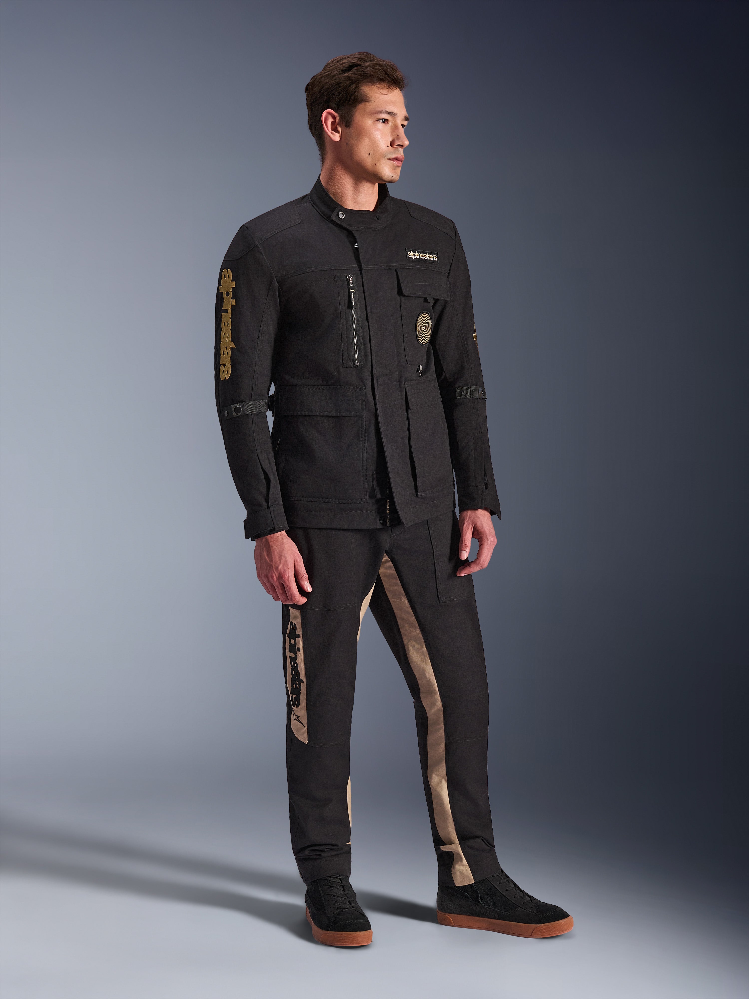Male model wearing Alpinestars Dice Jacket in Black, urban motorcycle jacket with vintage field jacket aesthetic, featuring multiple utility pockets and gold branding on the sleeve, paired with matching black and tan pants and black sneakers.