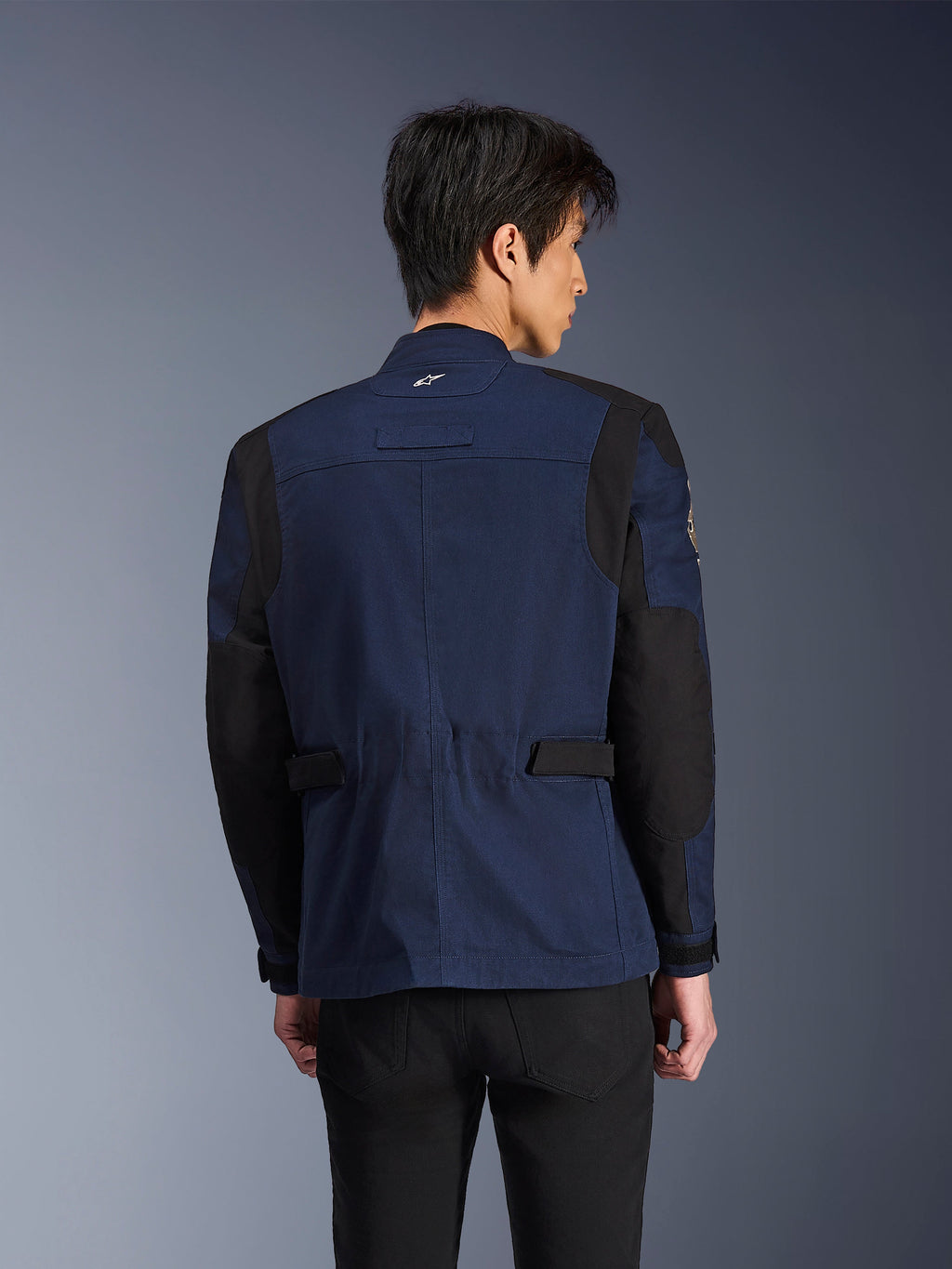 Alpinestars Dice Jacke, dunkelblaue und schwarze urbane Field-Motorradjacke, Rückansicht getragen von einem Mann, mit verstärkten schwarzen Schulter- und Ellbogenpartien und verstellbaren Taillenriegeln.