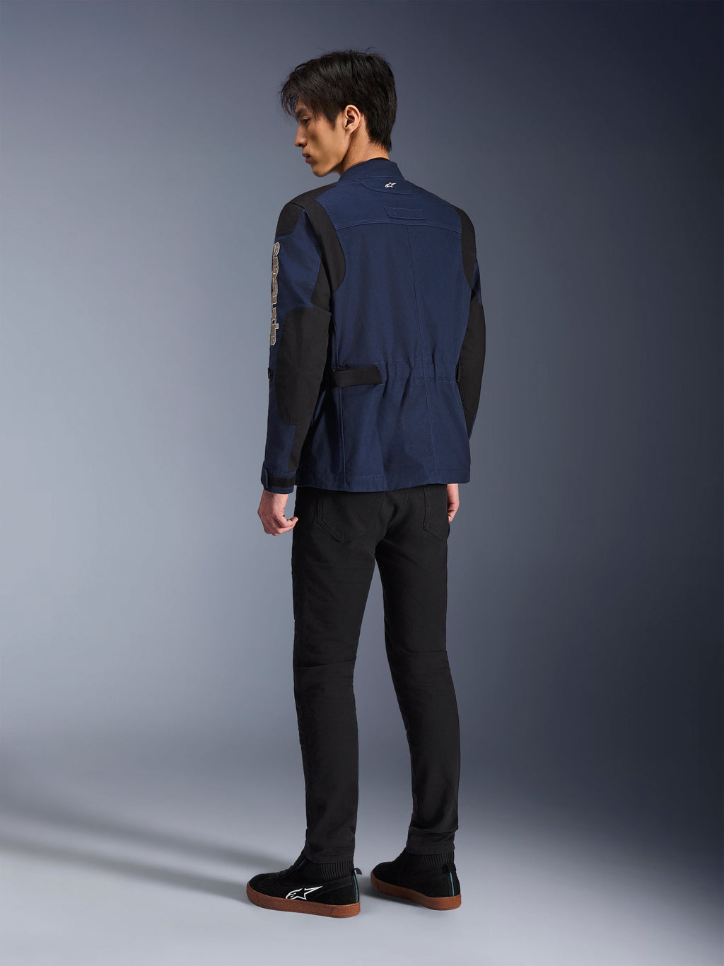 Männliches Model in der Rückansicht trägt die Alpinestars Dice Jacke in Dunkelblau und Schwarz, kombiniert mit einer schwarzen Hose und schwarzen Sneakern, urbane Motorrad-Fieldjacket mit Schulterfalten und verstellbaren Taillenriegeln.