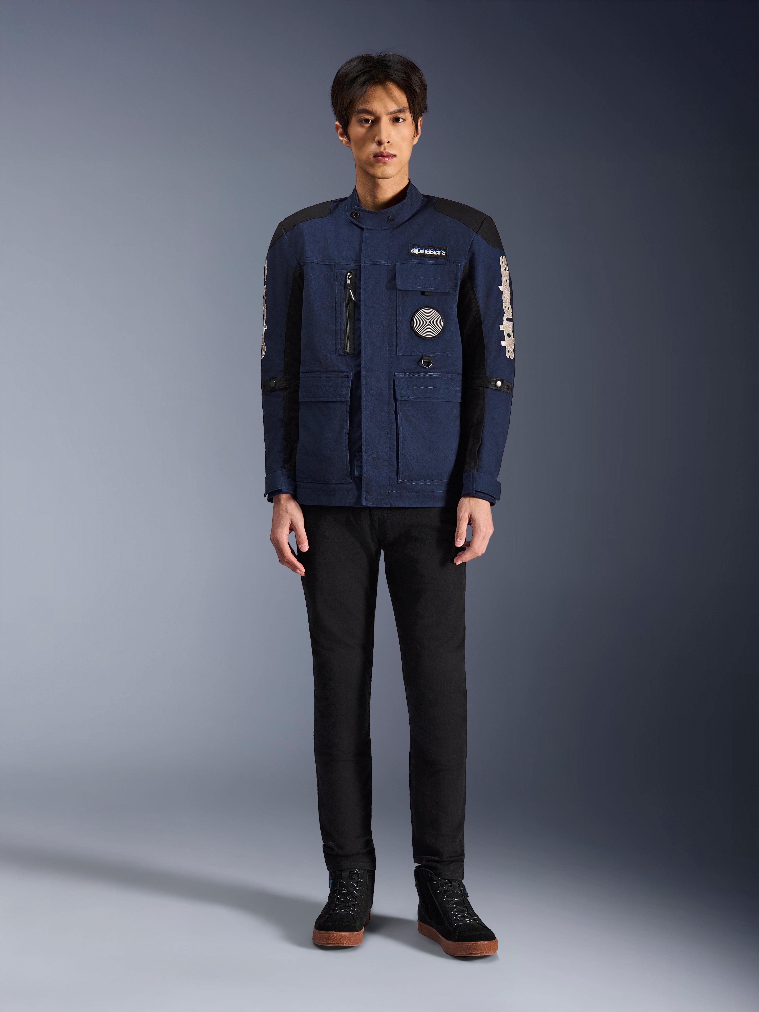 Alpinestars Dice Jacke, urbane Motorradjacke, Dunkelblau und Schwarz, getragen von einem Model mit schwarzer Hose und schwarzen Schuhen, modernes Fieldjacket-Design mit mehreren Taschen, vertikalem Brust-Reißverschluss und weißem Alpinestars-Branding auf den Ärmeln.
