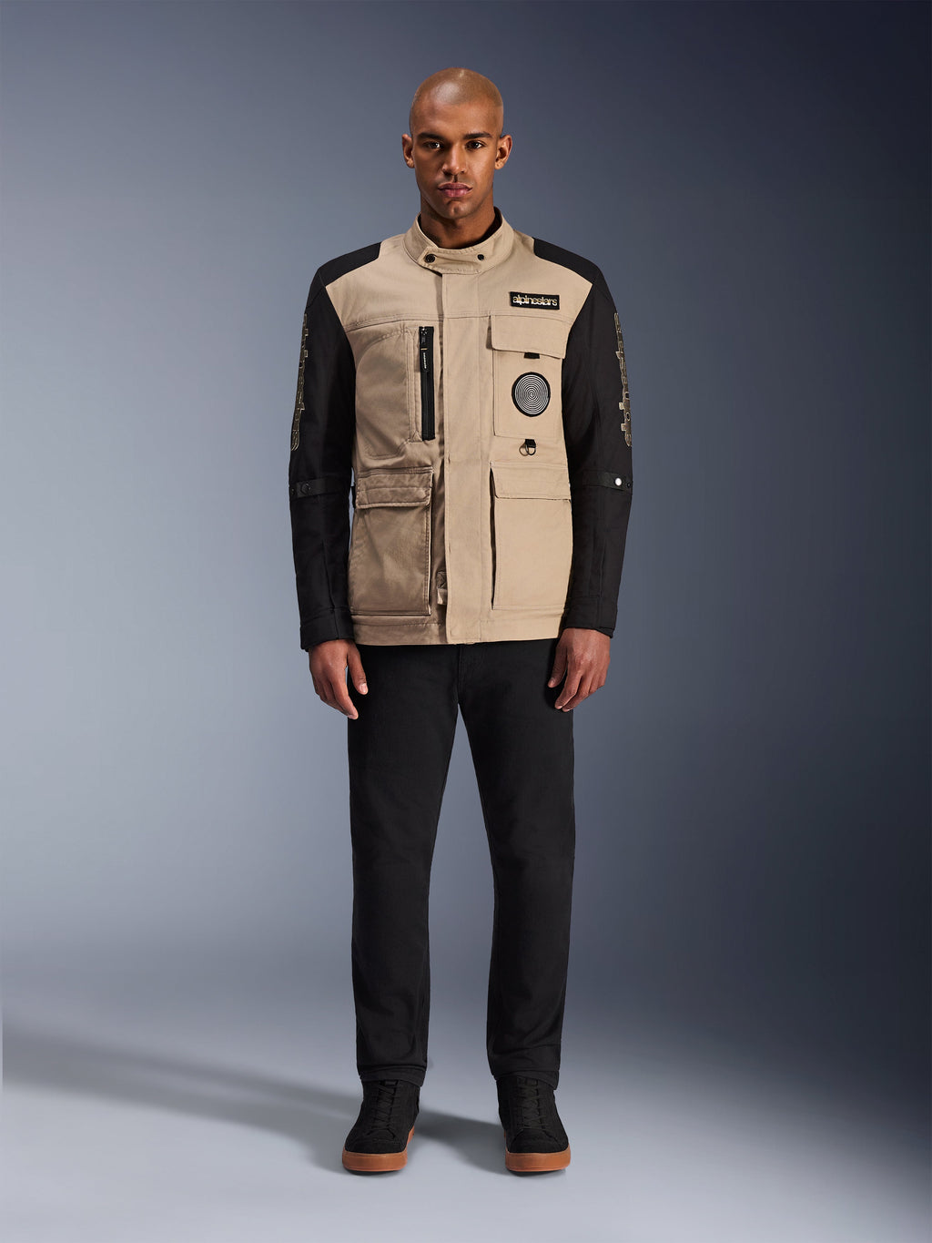 Alpinestars Dice Jacke, urbane Motorradjacke, Korpus in Aluminum-Beige mit schwarzen Ärmeln, von Arbeitskleidung inspirierte Silhouette mit vier funktionalen Fronttaschen, getragen mit schwarzer Hose und schwarzen High-Top-Sneakern, robuste straßentaugliche Fahrausrüstung