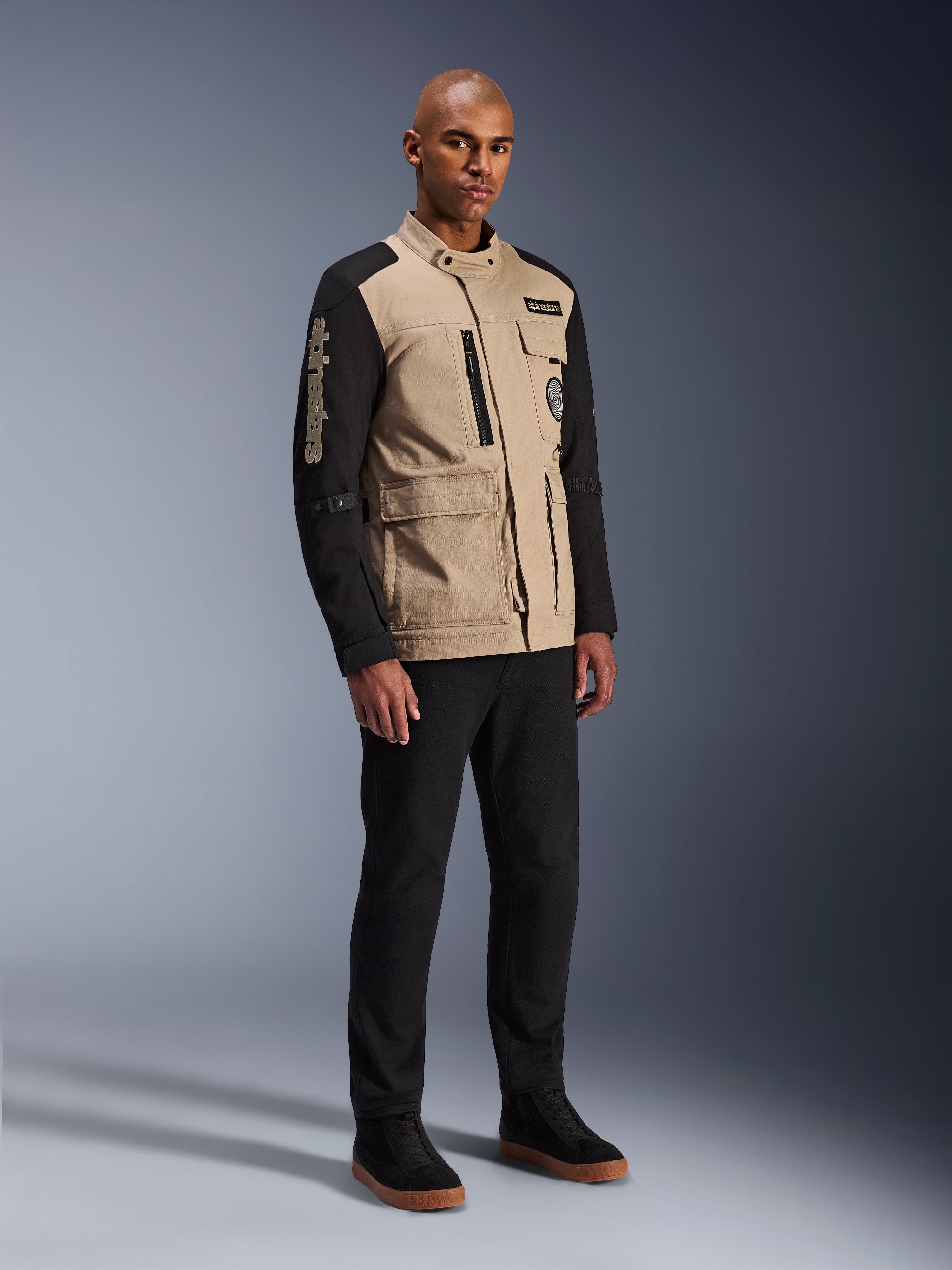 Alpinestars Dice Jacke, Urbane Motorradjacke, Aluminum Black und Tan, zweifarbiges Design mit schwarzen Ärmeln und khakifarbenem Torso, getragen von einem Model mit schwarzer Hose und schwarzen Sneakern, Workwear-inspirierte Ästhetik mit Utility-Taschen und gesticktem Branding auf dem Ärmel.