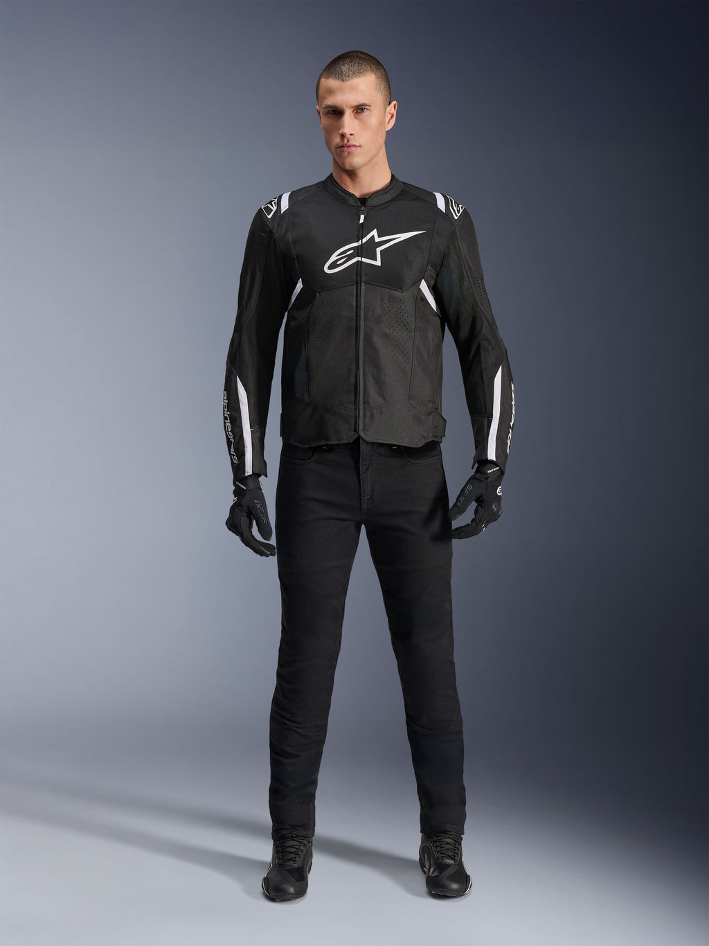 Alpinestars T-Sps Air V2 Jacke, schwarz-weiße Textil-Motorradjacke, mit großem weißem Brust-Logo und Mesh-Einsätzen, getragen von einem Model mit schwarzer Ausrüstung für sportliches Fahren in der Stadt
