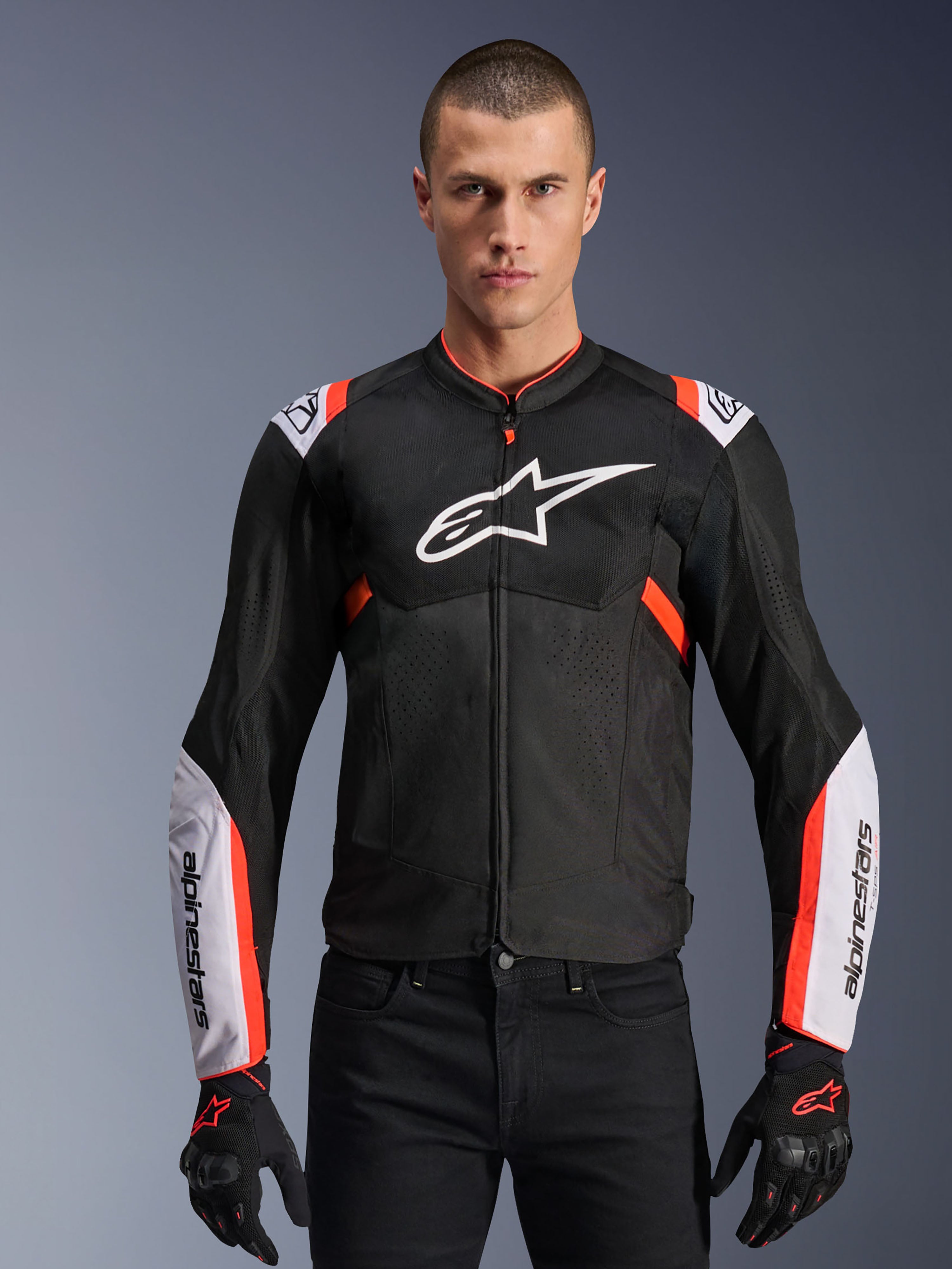 Alpinestars T-Sps Air V2 Jacke, Schwarz mit weißen und leuchtend roten Akzenten, Motorrad-Sportjacke, Mann trägt ein Design mit Mesh-Belüftung, großem Brustlogo und Ärmeln mit Markenschriftzug, kombiniert mit schwarzen Handschuhen und Hosen
