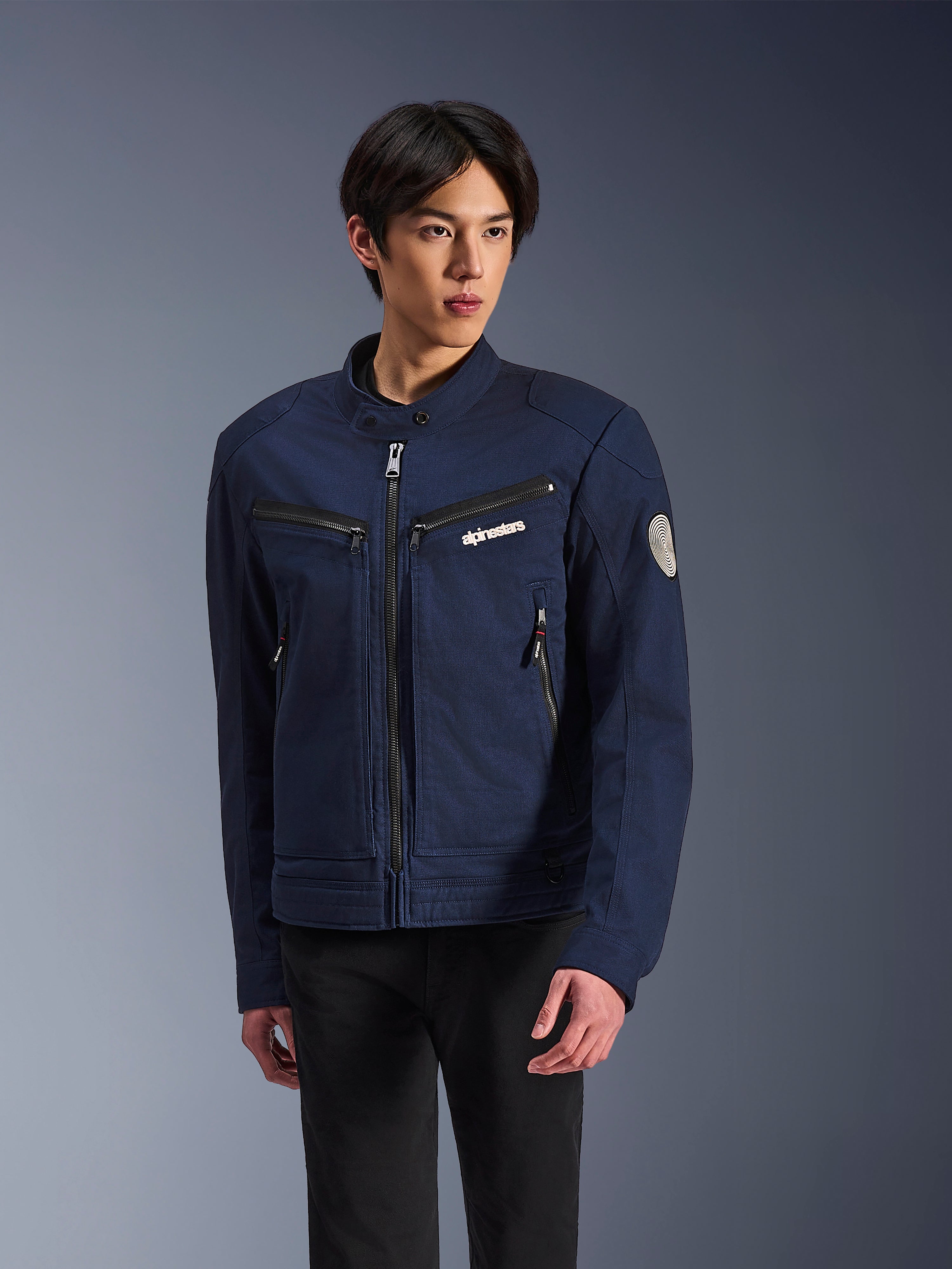 Alpinestars Bruiser Jacke, dunkelblaue Textil-Motorradjacke, getragen von einem Mann mit schwarzer Hose, mit Reißverschlusstaschen, weißem Alpinestars-Brustlogo und rundem Oscar-Spiral-Aufnäher am Ärmel, urbaner Streetwear-Stil für Fahrer