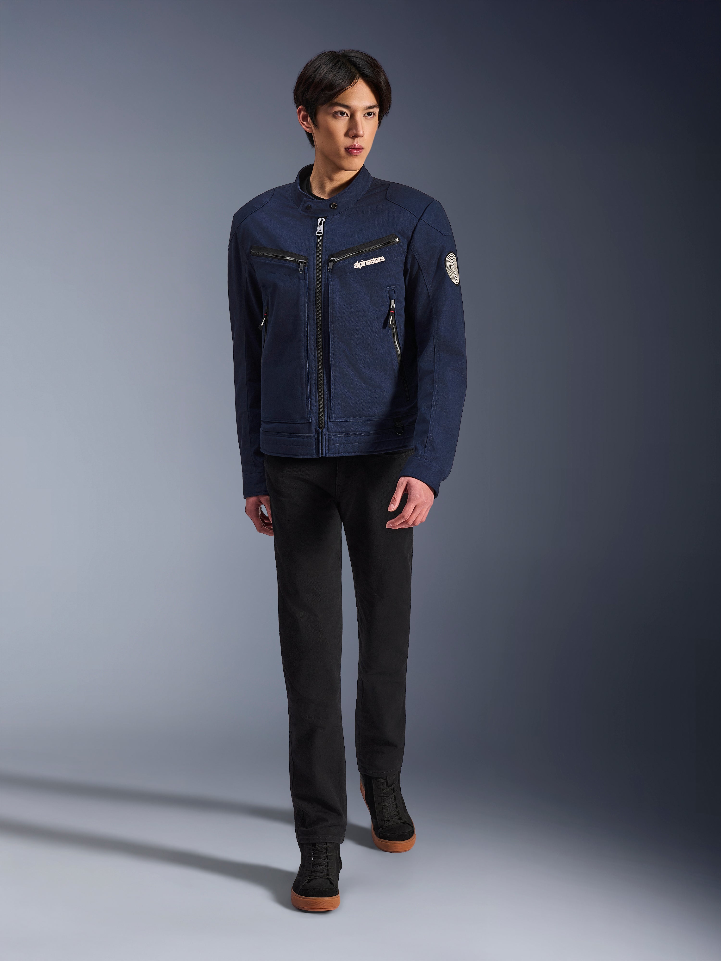 Männliches Model in Alpinestars Bruiser Jacke in Dark Blue, urbane Textil-Motorradjacke, kombiniert mit schwarzer Hose und schwarzen Sneakern, Workwear-inspirierte Ästhetik mit Reißverschlusstaschen und rundem Ärmel-Patch, zeitgemäße Streetwear für das Motorradfahren.
