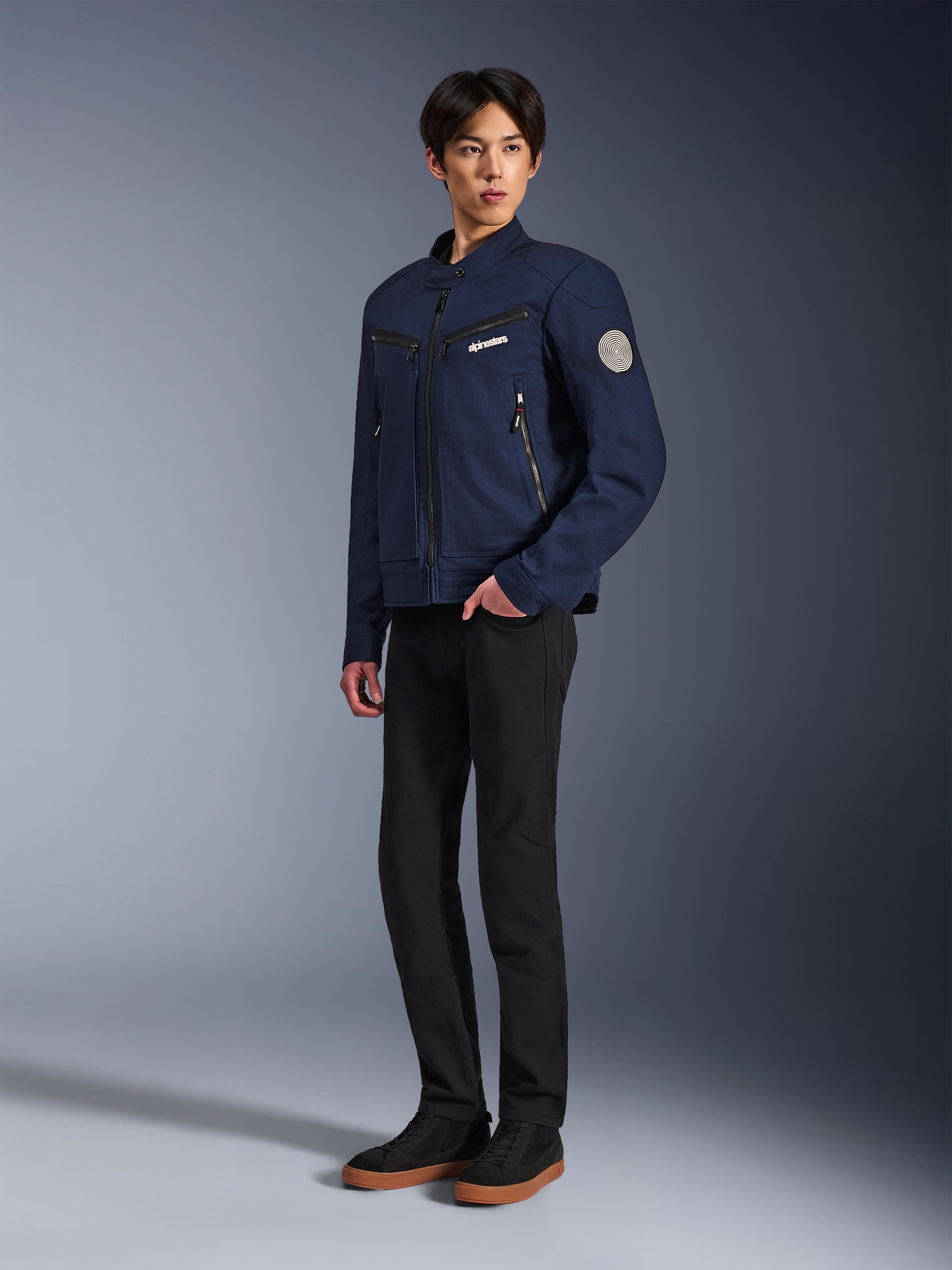 Alpinestars Bruiser Jacke in Dunkelblau, urbane Textil-Motorradjacke mit von Workwear inspiriertem Design, mit Reißverschlusstaschen an der Brust und Oscar Spiral-Ärmel-Patch, getragen mit schwarzer Hose und lässigen Sneakern.