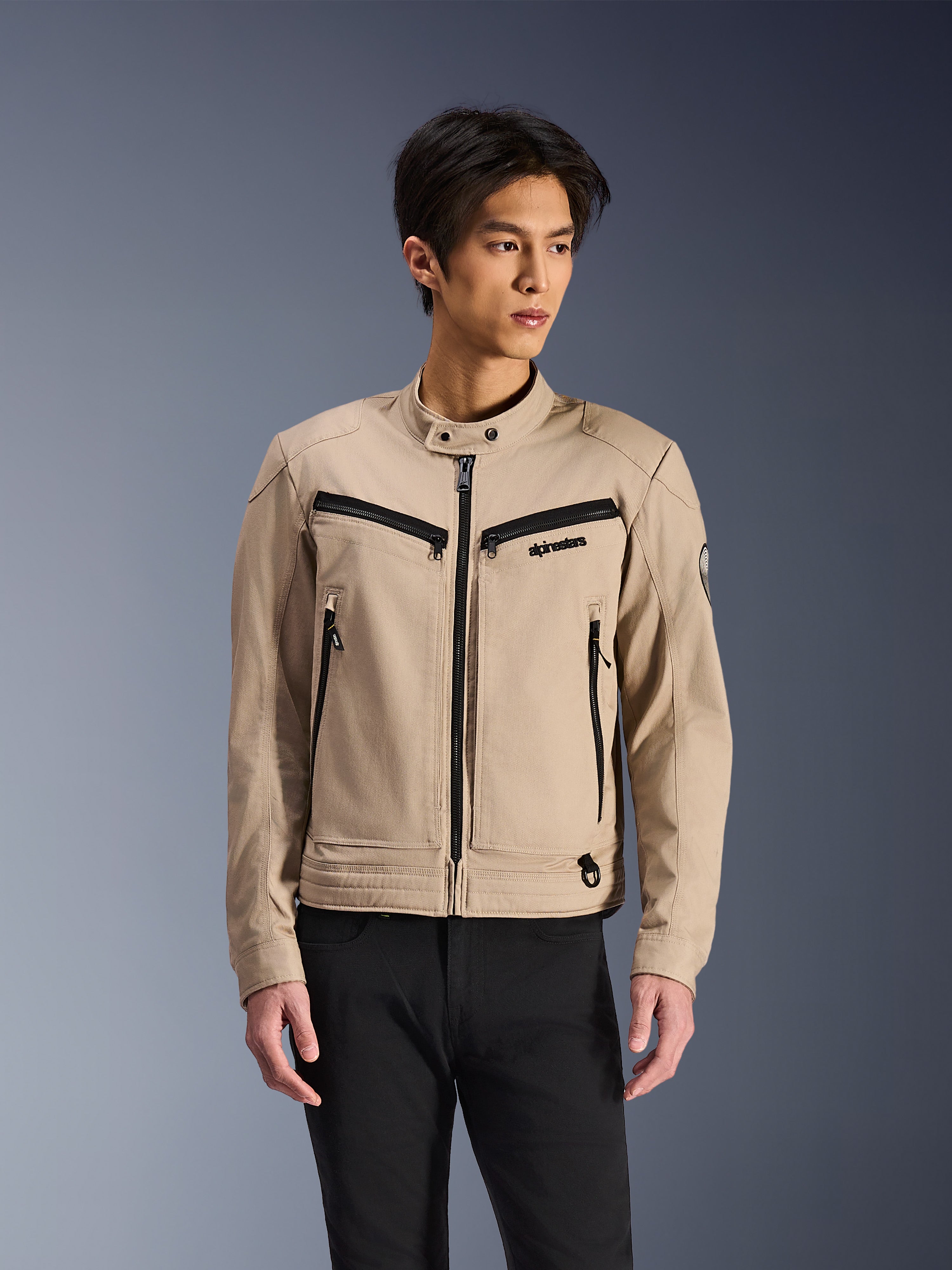 Alpinestars Bruiser Jacke, beige Motorradjacke getragen von einem männlichen Model, schwarze Reißverschlussdetails an Brust- und Seitentaschen, hoher Druckknopfkragen, Workwear-inspirierte urbane Motorradbekleidung, kombiniert mit einer schwarzen Hose