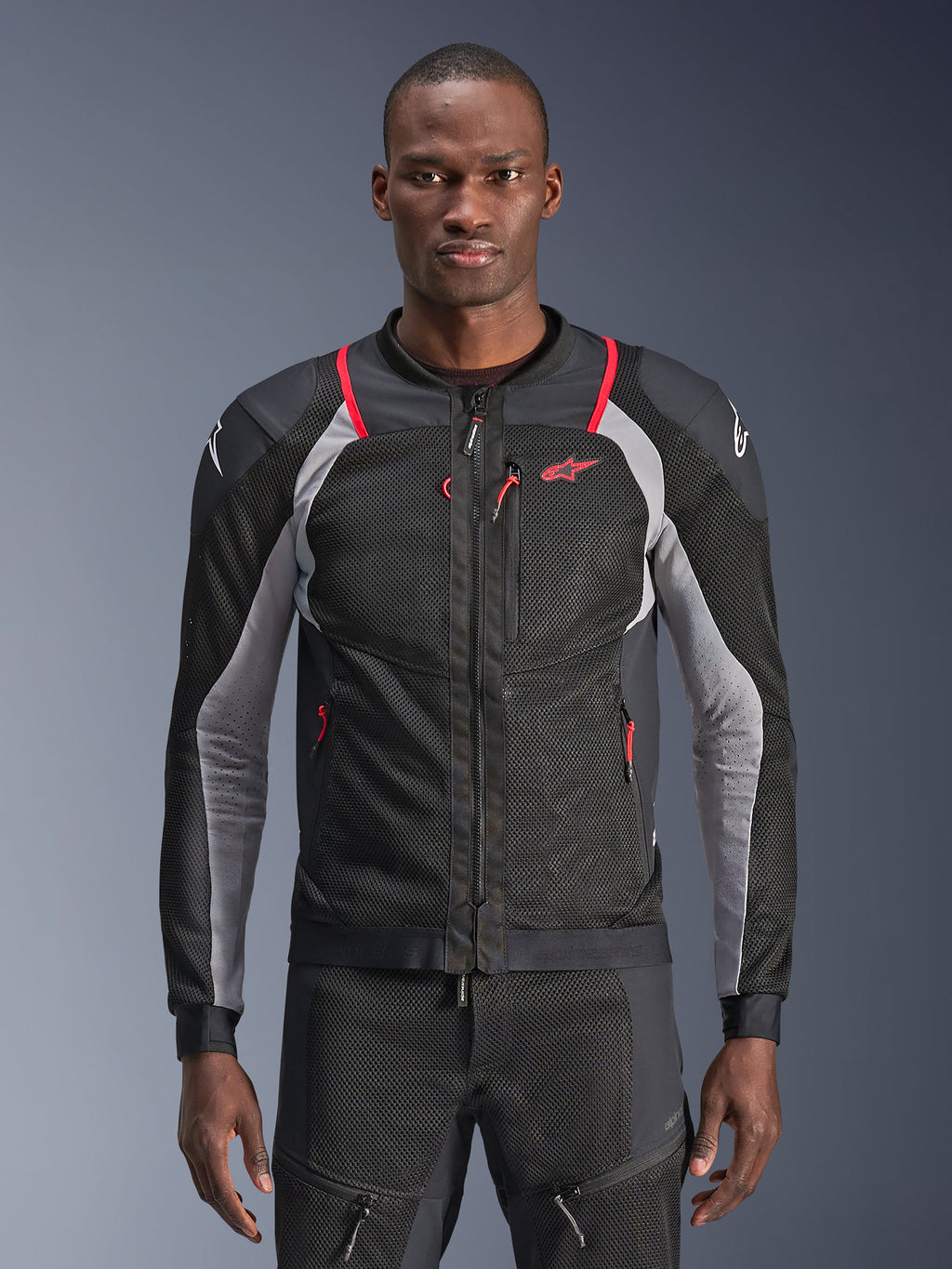 Alpinestars Troop-Air Pro Jacke, hochwertige gepanzerte Air-Jacke, Schwarz, Ash Gray und Bright Red, technische Mesh-Konstruktion mit roten Akzenten an den Schultern und Reißverschlüssen, graue Details an den Ärmeln und Alpinestars-Logo auf der Brust und den Schultern, getragen von einem Model vor grauem Hintergrund.
