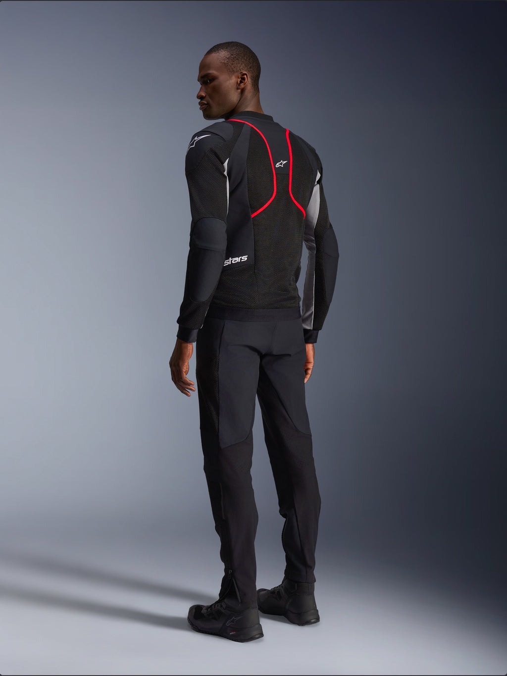 Alpinestars Troop-Air Pro Jacke, Motorradjacke, Schwarz, Eschegrau und Hellrot, Rückansicht am Modell, mit roten Akzentlinien und Mesh-Belüftungseinsätzen, getragen mit schwarzer Hose und Stiefeln, urbaner Motorradstil
